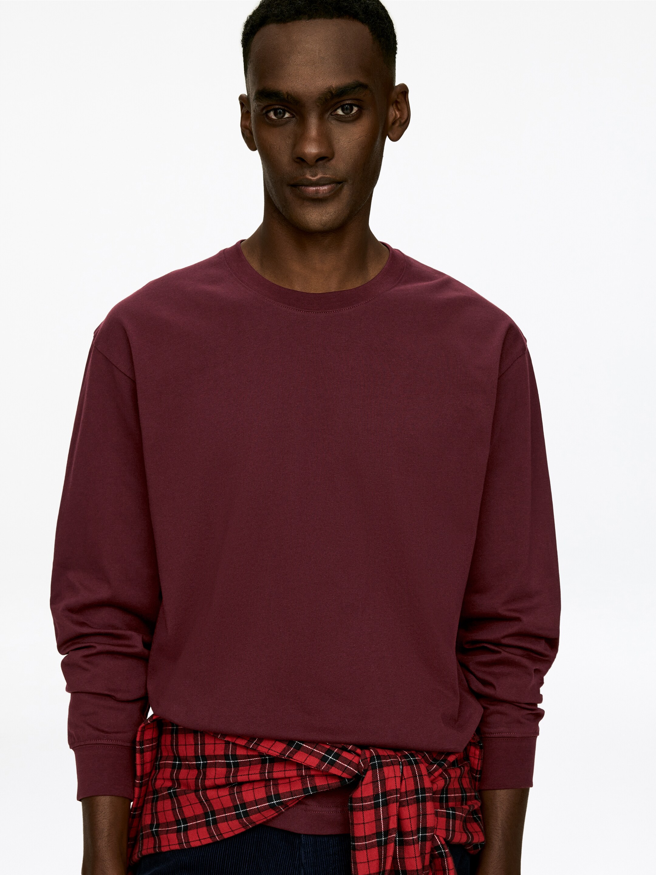 Agrandir l'image: La chemise bordeaux à manches longues présente une texture lisse, un col ras du cou et des poignets côtelés, tournée vers l'avant. Une chemise à carreaux rouge et noire est nouée autour de sa taille.