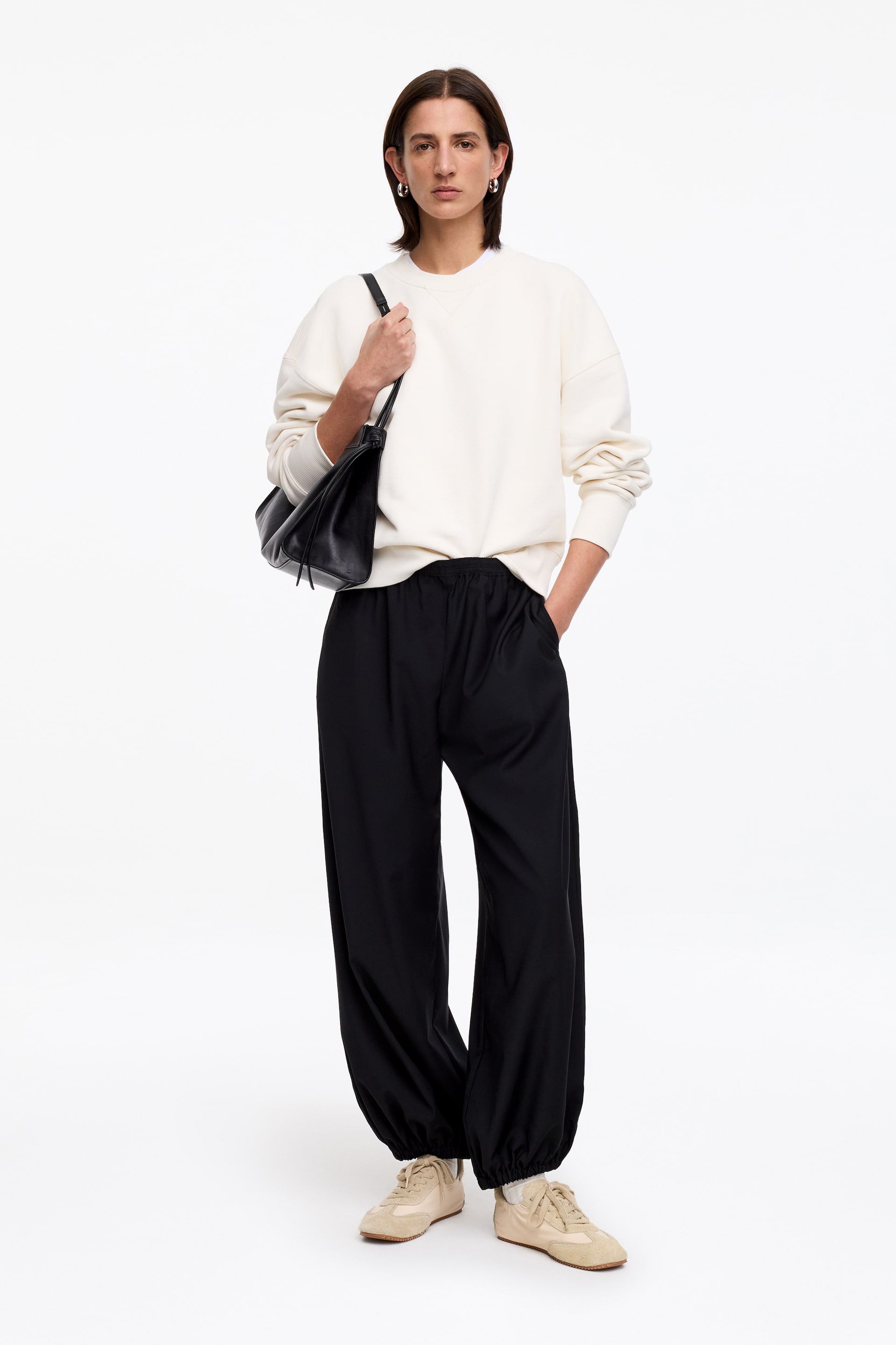 Ver imagen más grande: Zapatillas de inspiración retro, confeccionadas en ante beige y nylon con detalles de pespuntes, son llevadas por la modelo. Cuentan con cierre de cordones, y los pantalones negros de la modelo, con tobillos elásticos, descansan sobre ellas, revelando calcetines blancos.