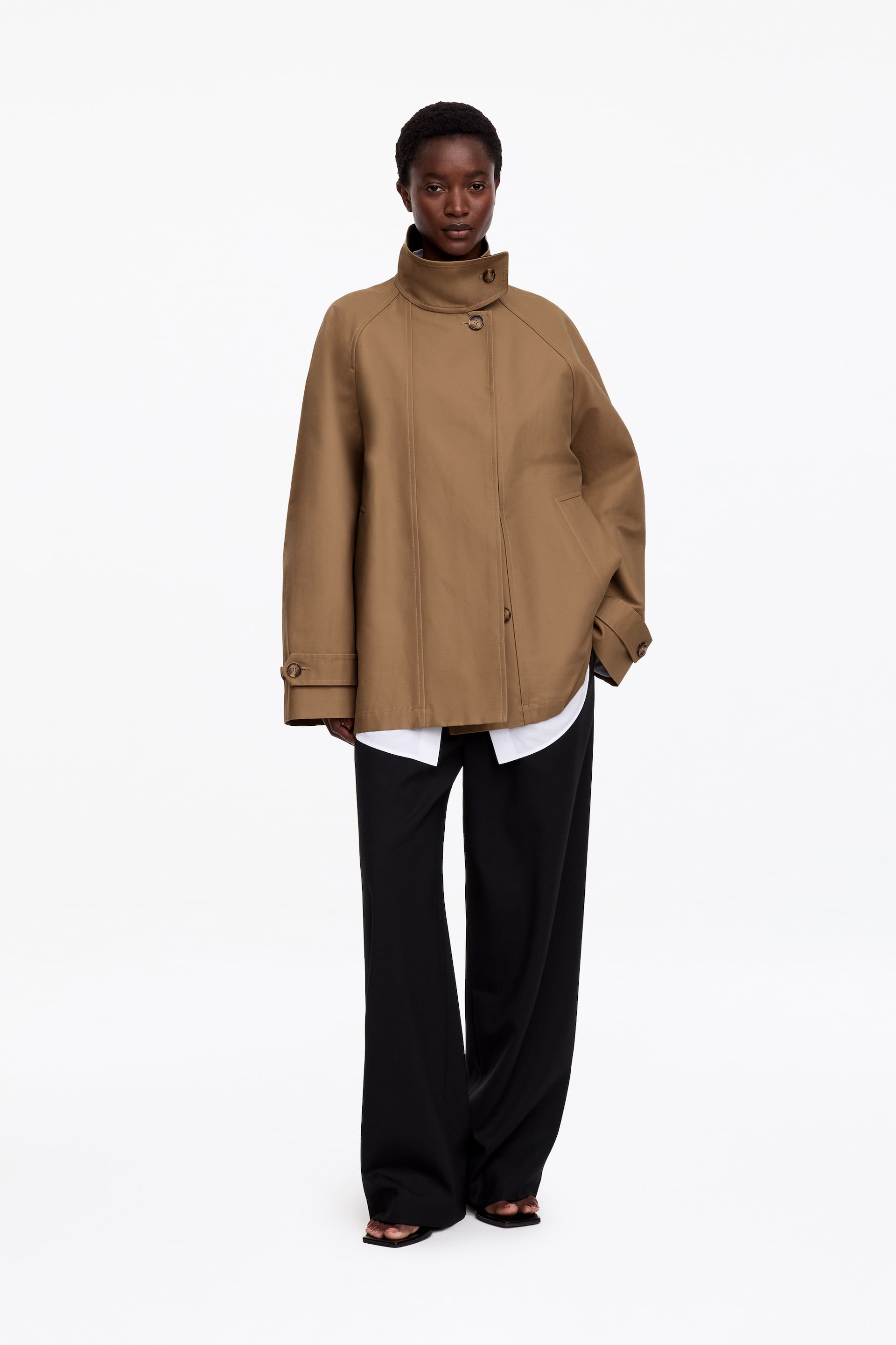 Visualizza immagine più grande: La modella indossa una giacca in cotone color beige con collo alto a imbuto, spalle raglan e alamari con bottoni sui polsini. La giacca è indossata sopra una camicia bianca con bottoni e pantaloni neri a gamba larga.