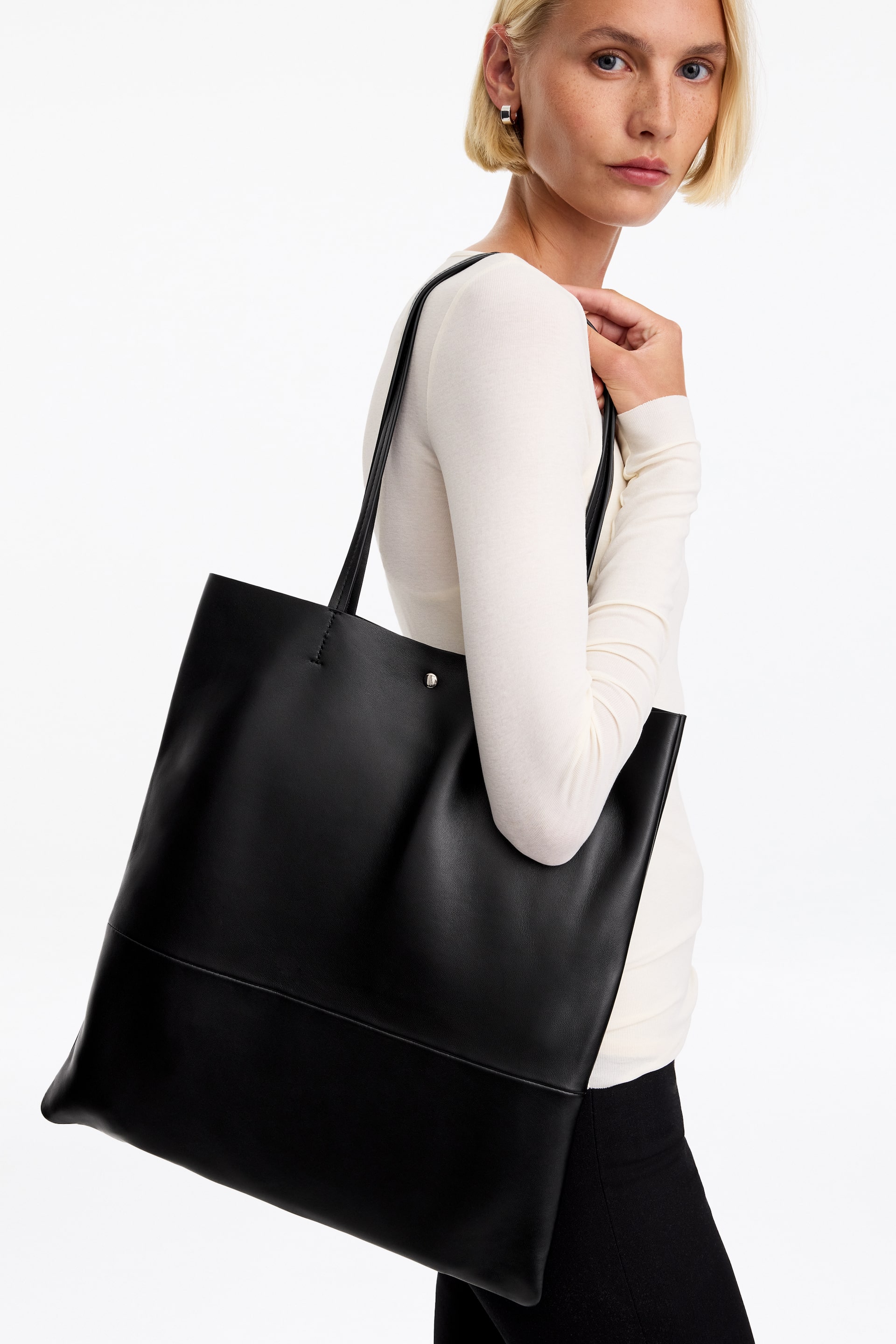 Agrandir l'image: Un sac cabas en cuir noir au design minimaliste, doté de fines anses d’épaule et d’une fermeture à bouton-pression argentée, est porté à l’épaule par le mannequin. Le mannequin tient la bandoulière d’une main tout en portant un haut crème à manches longues.