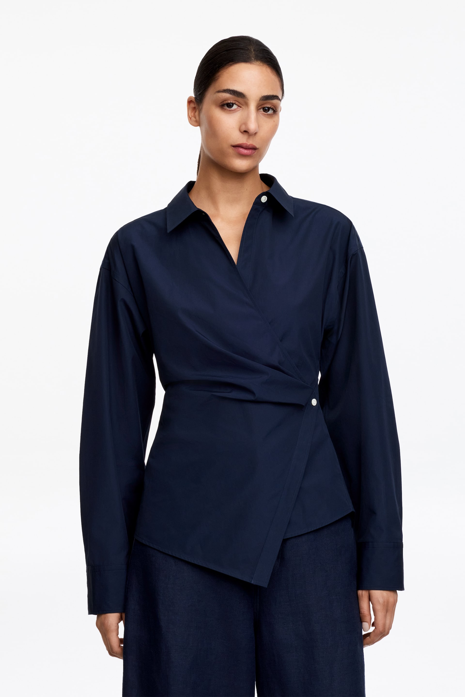 Grotere afbeelding bekijken: Een marineblauwe overslagblouse is frontaal gericht, met een puntige kraag en lange mouwen met knoopmanchetten. De asymmetrische overslag aan de voorkant wordt vastgezet door een enkele witte knoop aan de rechterkant.