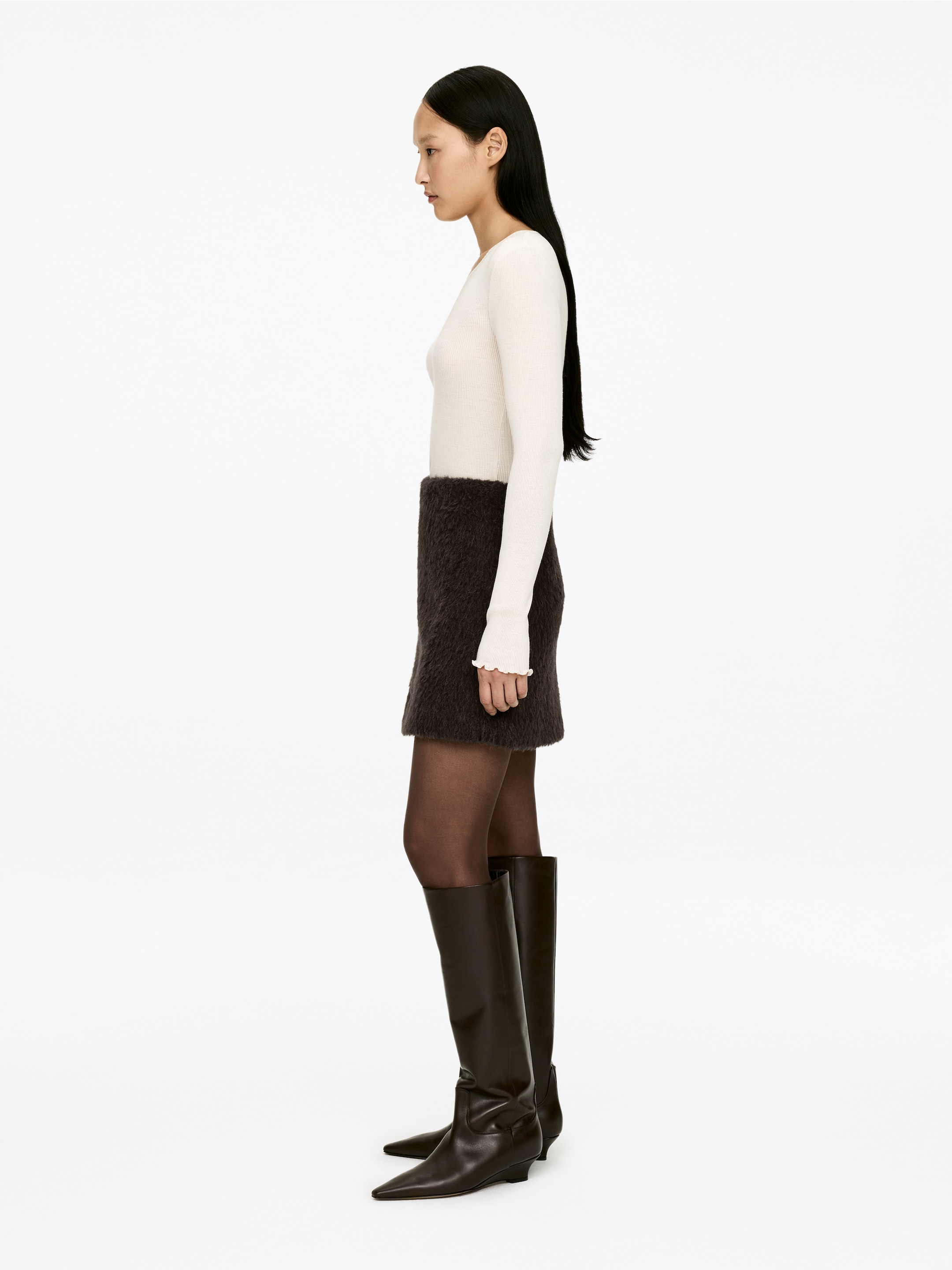 Agrandir l'image: Un ensemble comprenant un haut côtelé à manches longues blanc cassé, une mini-jupe duveteuse marron foncé, des bas sombres transparents et des bottes marron foncé montant jusqu'au genou est présenté en profil latéral. Le haut présente des poignets à volants et les bottes arborent des bouts pointus et des talons bas.
