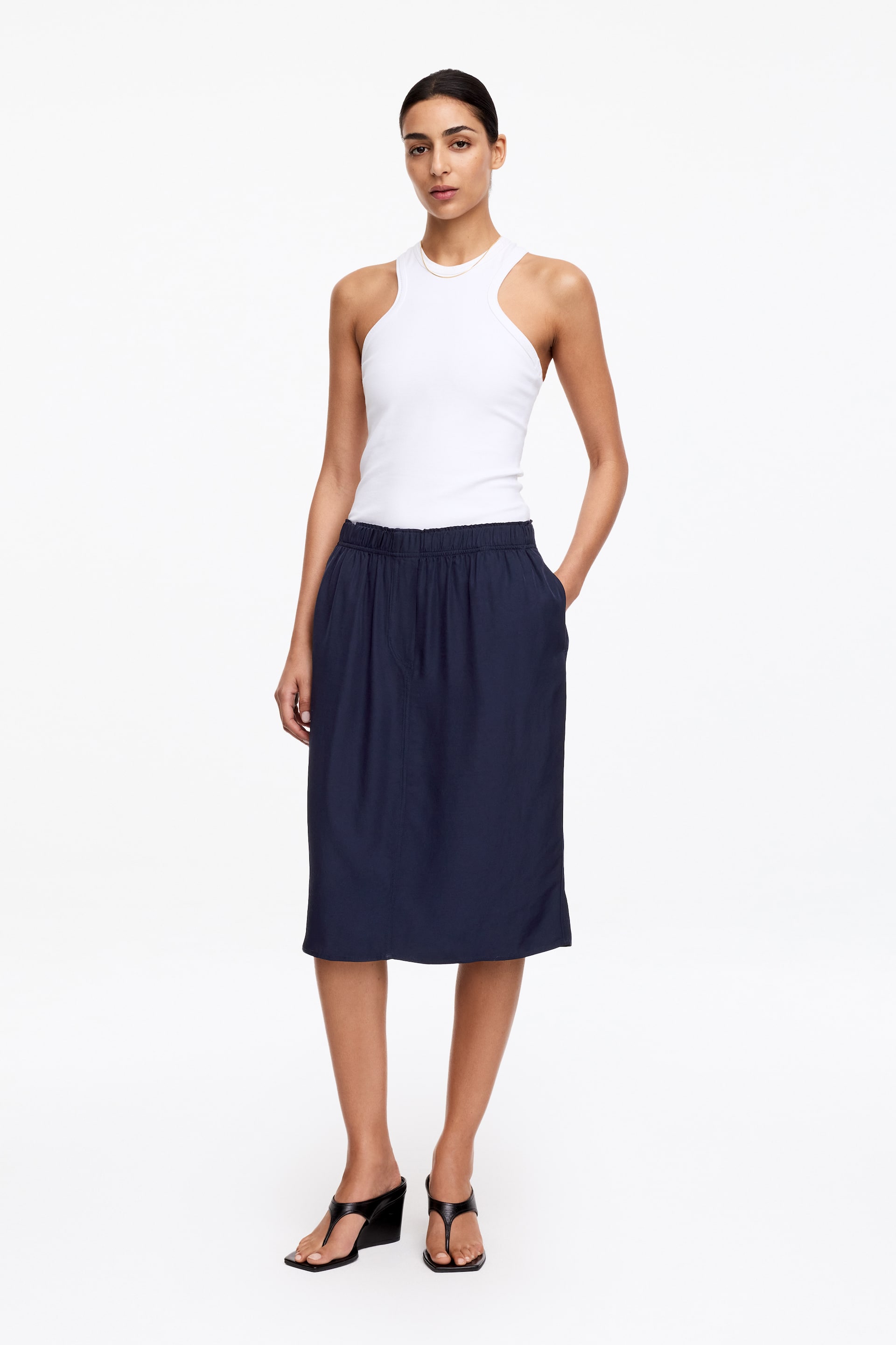 Se større billede: En hvid ribstrikket tanktop med høj crew neck og indskårne skuldre. Den er stukket ned i en marineblå midinederdel, som har en elastisk talje og sidelommer.
