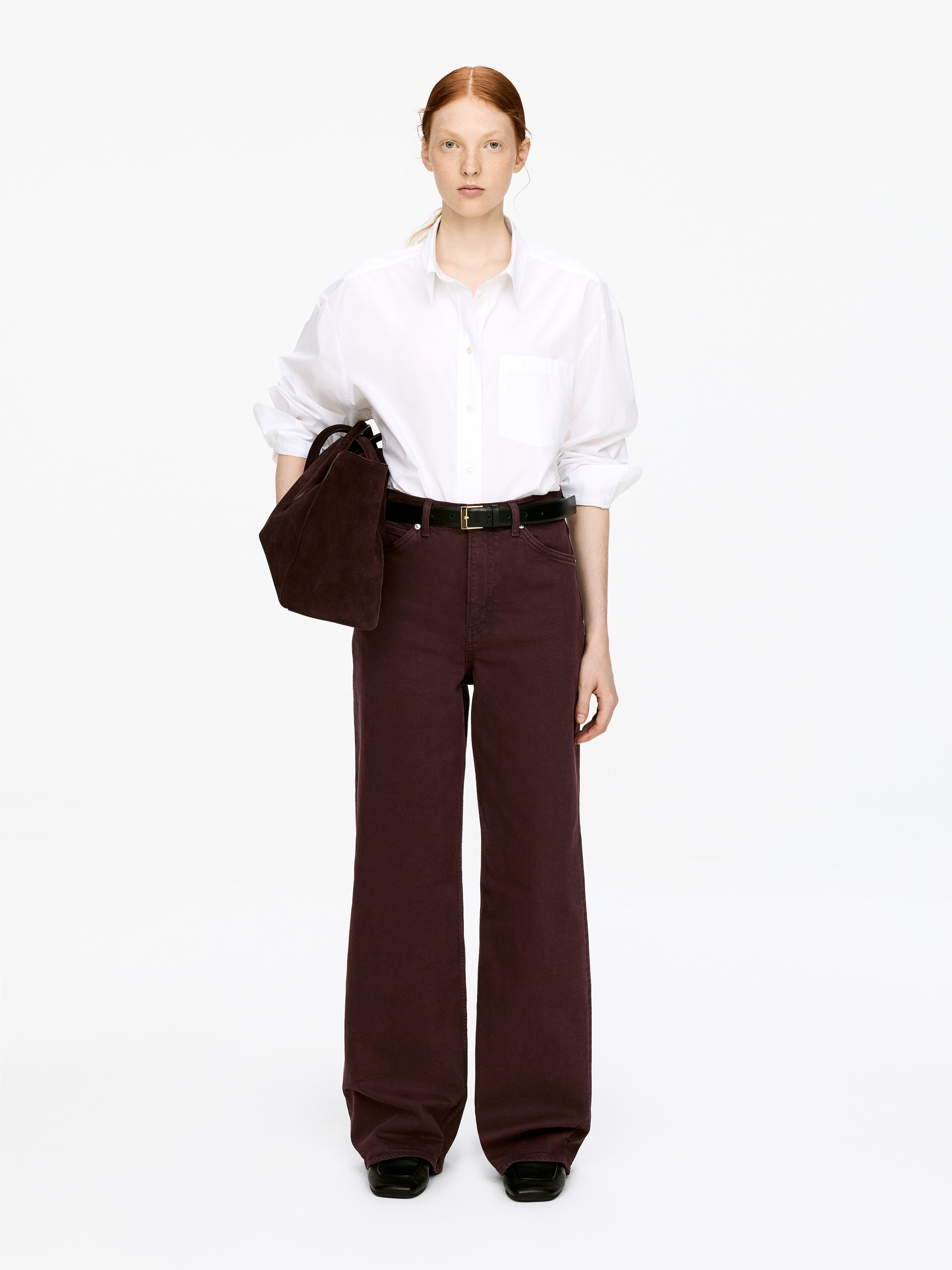 Visualizza immagine più grande: Pantaloni di jeans elasticizzati color borgogna scuro con vita alta, vestibilità aderente e gambe svasate, indossati dalla modella. Una cintura nera stringe i pantaloni in vita e una camicia bianca è infilata dentro.
