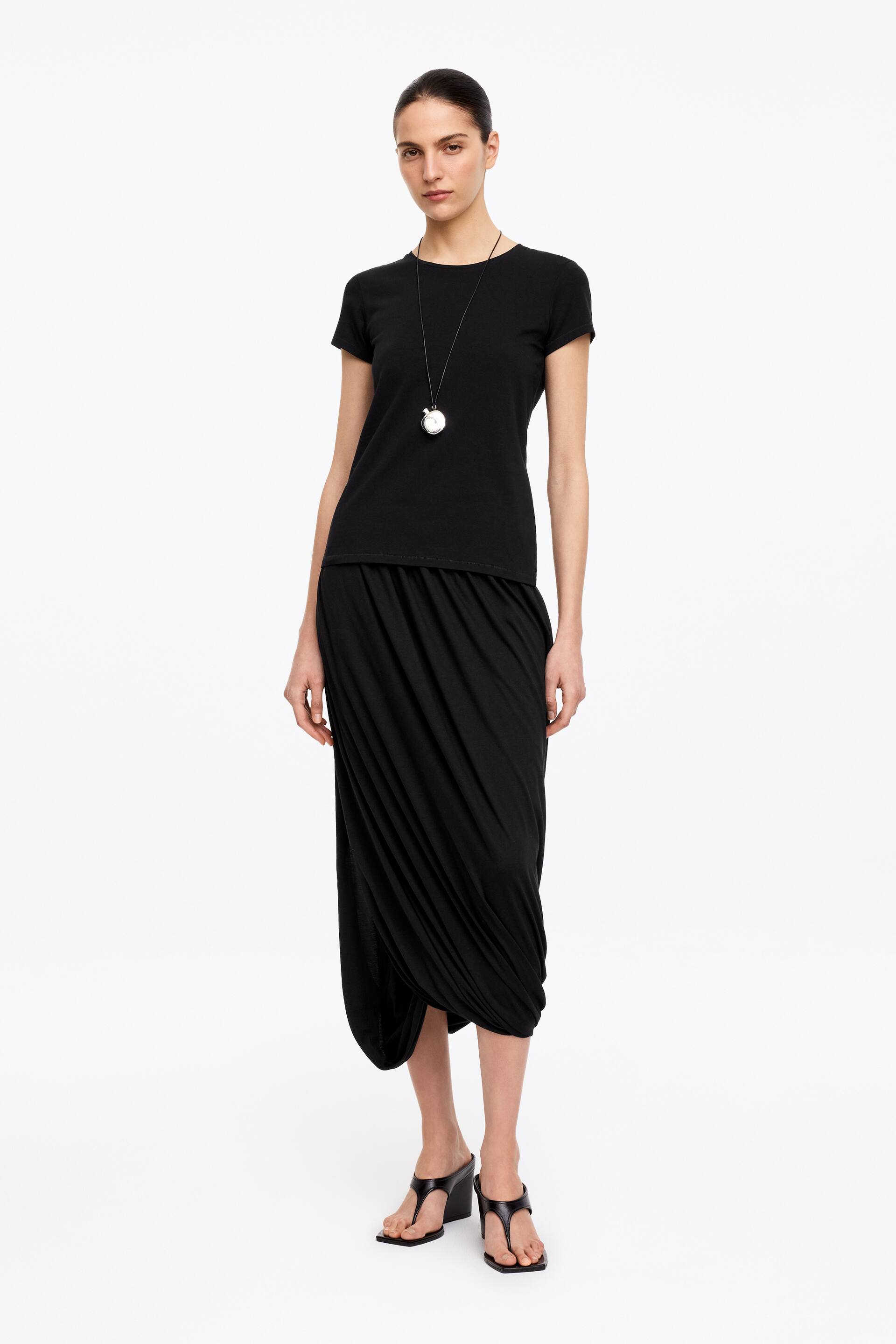 Ver imagem maior: Uma t-shirt preta de manga curta e gola redonda está a ser usada com uma saia midi preta, com drapeados assimétricos e franzidos no lado esquerdo. Um colar comprido preto com um pendente circular em prata está a complementar a t-shirt, e sandálias pretas de cunha com tiras completam o visual.