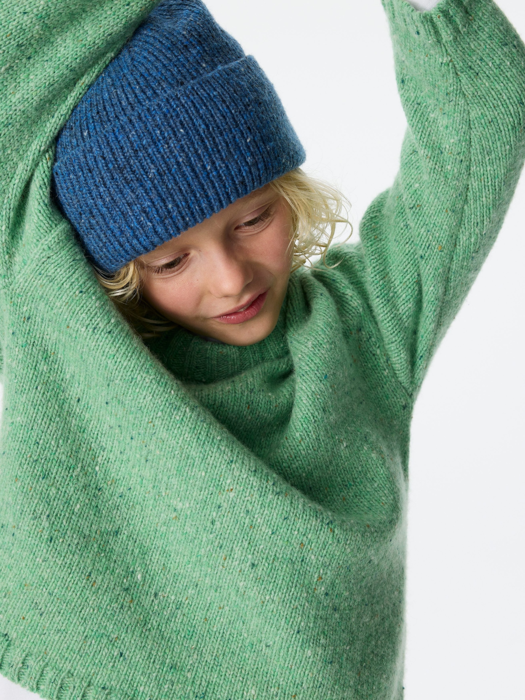 Größeres Bild anzeigen: Ein hellgrüner gesprenkelter Strickpullover wird getragen, mit hochgeschobenen Ärmeln, die die Textur des Kleidungsstücks und den gerippten Kragen zur Schau stellen, während eine blaue gerippte Strickmütze den Kopf bedeckt.