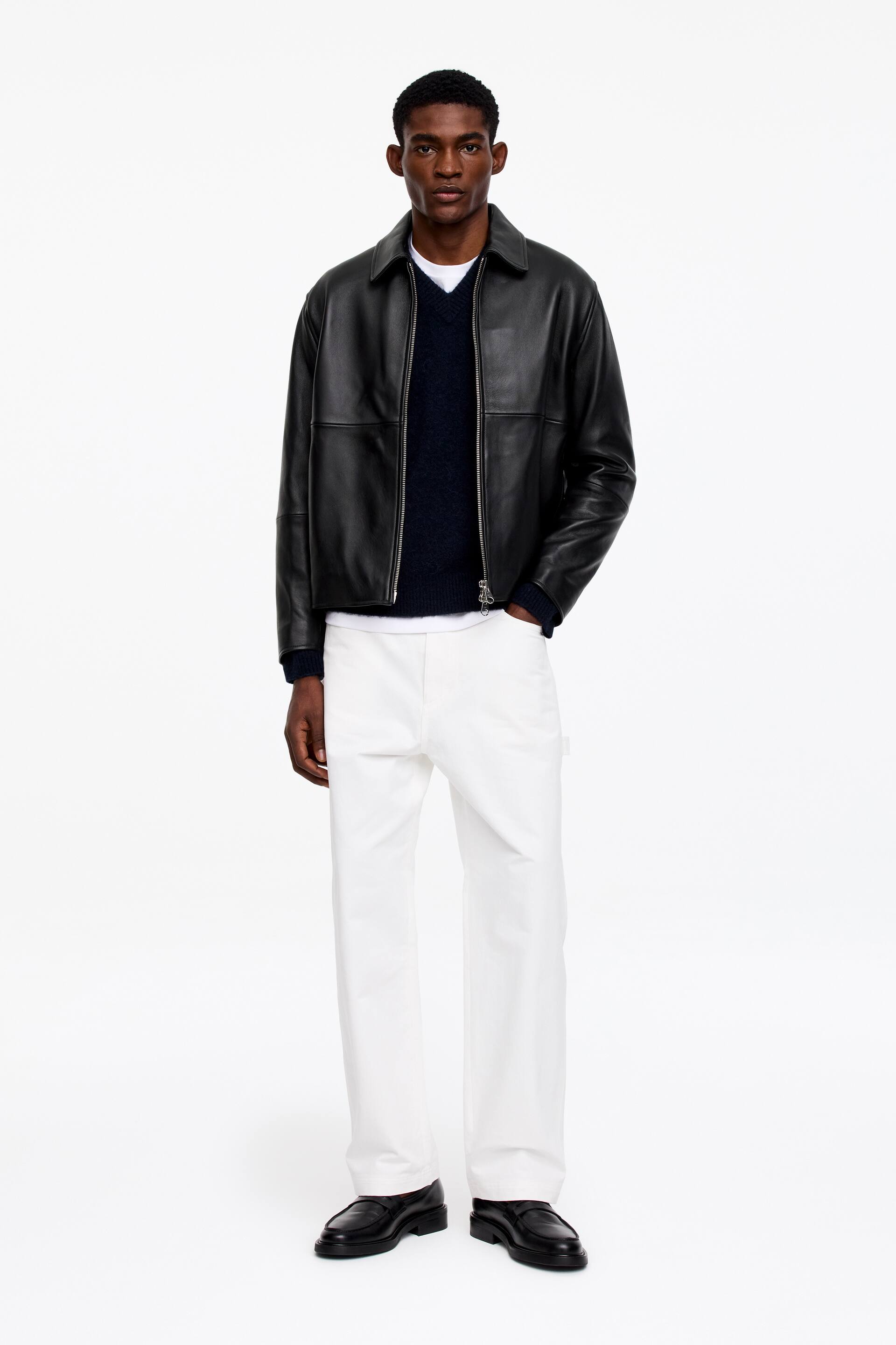 Vezi imaginea mai mare: O geacă bomber din piele neagră, cu guler ascuțit și fermoar argintiu pe toată lungimea, este purtată deschis peste un pulover bleumarin cu decolteu în V și un tricou alb cu guler rotund. Pantaloni albi cu croială dreaptă și buzunare laterale sunt purtați, asortați cu mocasini din piele neagră cu talpă groasă.