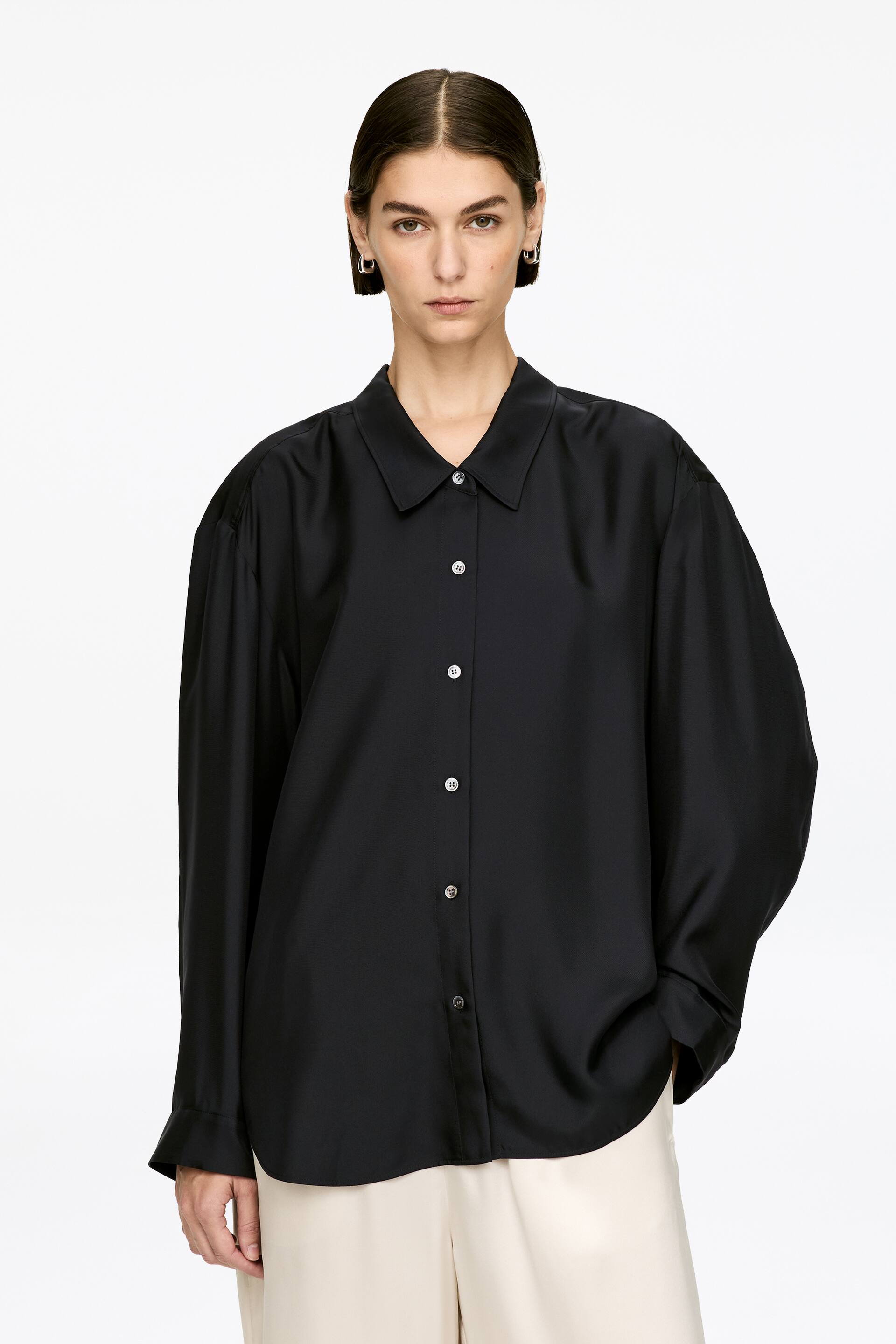 Größeres Bild anzeigen: Ein schwarzes Hemd, gefertigt aus reiner Seide und gestaltet mit einem niedrigen Kragen, Knopfverschluss und einem abgerundeten Saum, wird vom Model getragen. Das Model steht, mit einer Hand in der Tasche einer hellen Hose, und der Stoff des Hemdes fällt locker.