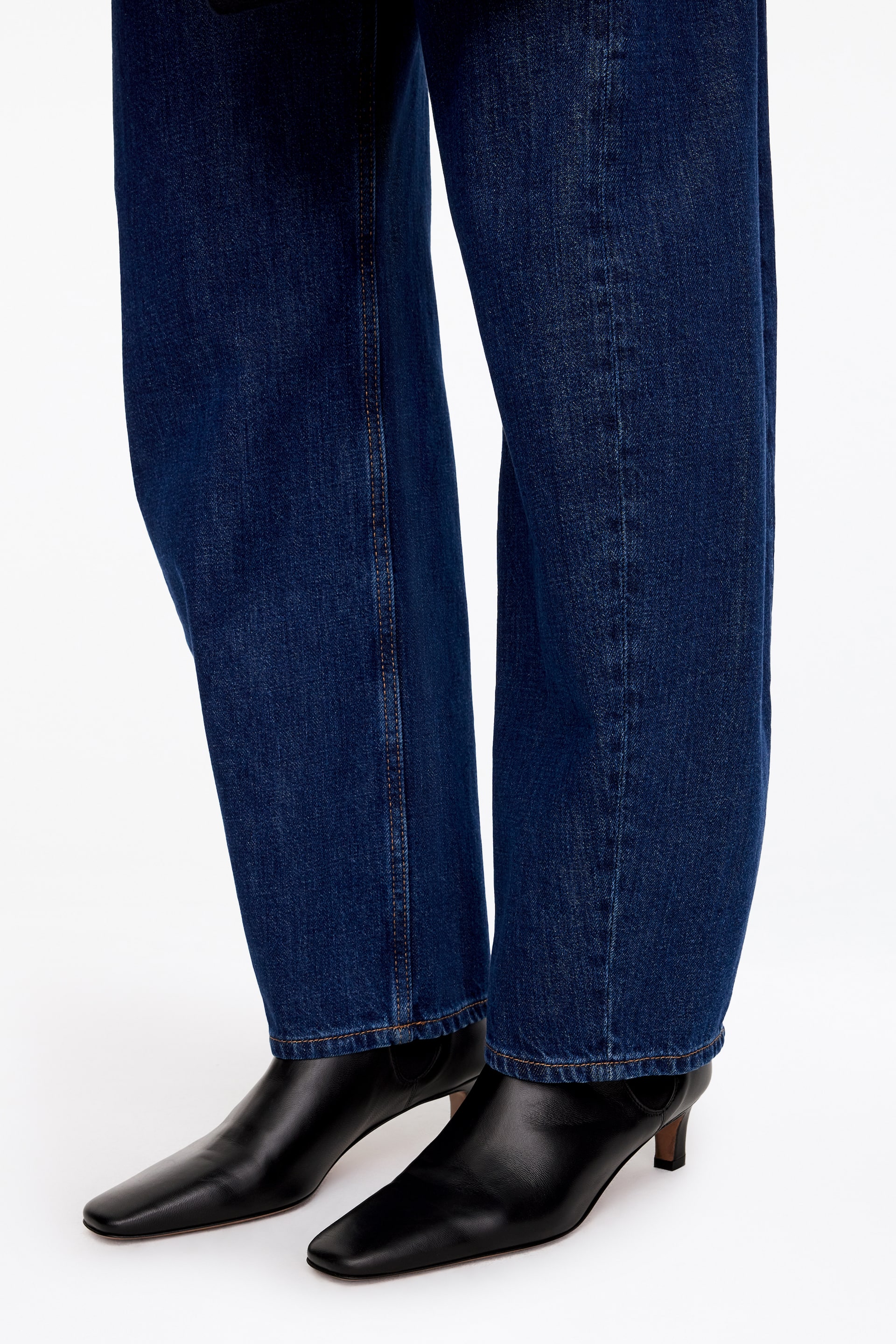 Agrandir l'image: Des bottines noires en cuir, à la silhouette allongée, dotées d'un bout carré souple et d'un petit talon, sont portées par le mannequin. Les bottines sont associées à un jean en denim bleu foncé à jambe large.