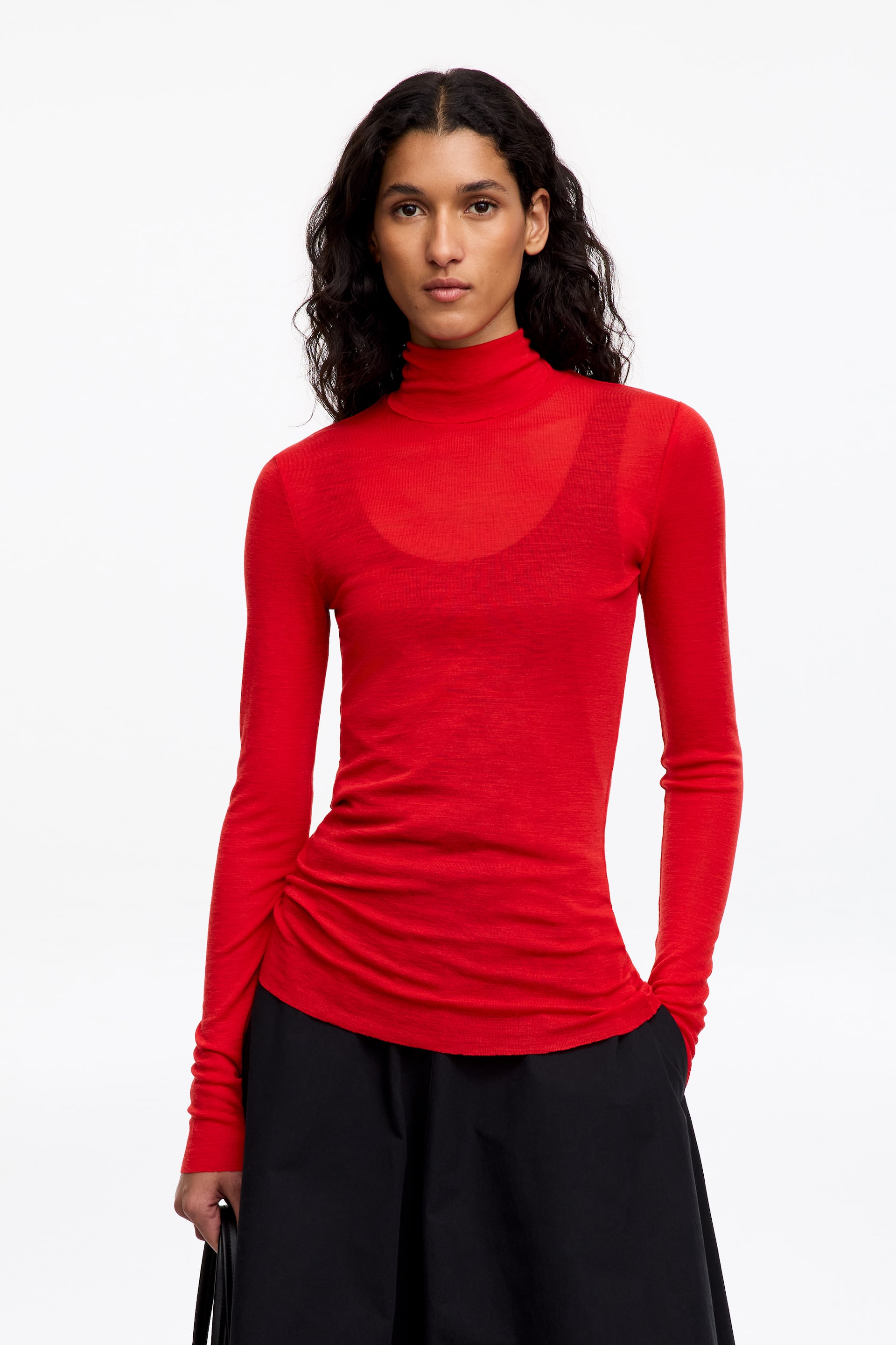 Größeres Bild anzeigen: Das Model trägt ein rotes, halbtransparentes Rollkragenoberteil mit langen Ärmeln, das über einem passenden roten Tanktop getragen wird. Das fein gestrickte Oberteil ist mit einer schwarzen Hose kombiniert, wobei das Model eine Hand lässig in der Tasche hält.