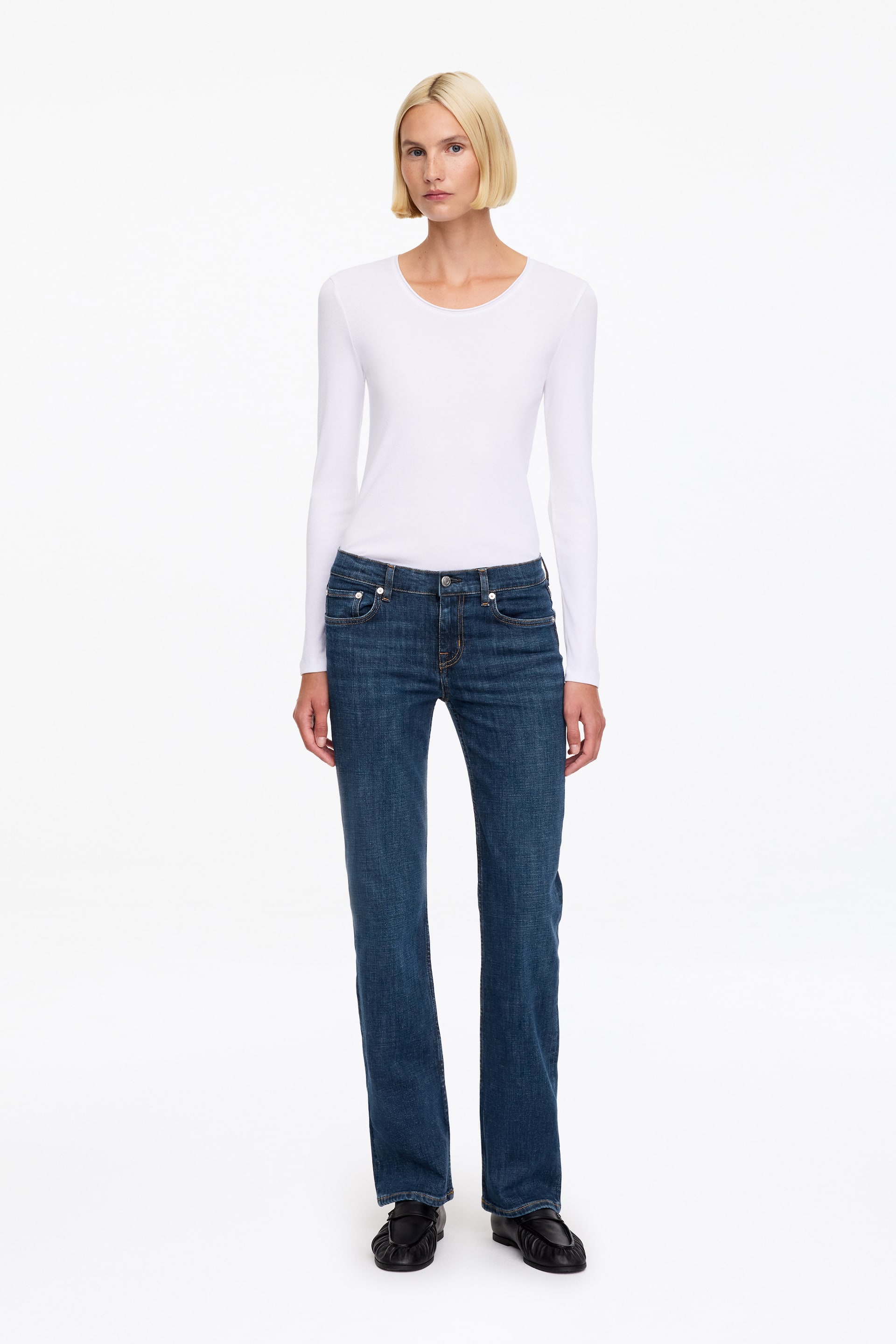 Ver imagen más grande: Unos vaqueros de denim con lavado azul oscuro, de tiro bajo, corte slim y pernera recta, lucidos por la modelo. Los vaqueros se combinan con una camisa blanca de manga larga metida por dentro y zapatos negros.