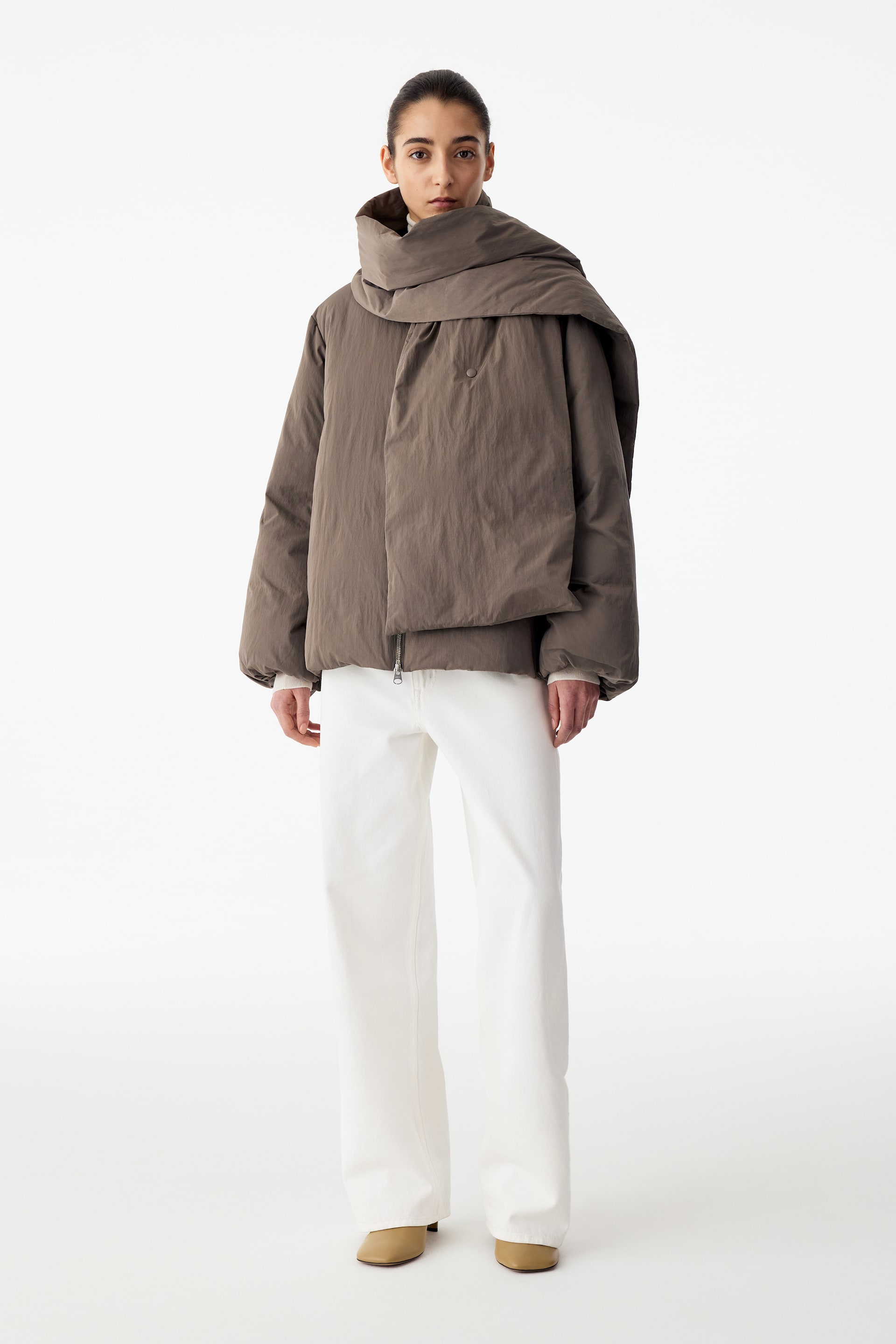 View larger image: An upcycled down jacket in a muted brown colour is being worn by the model, featuring a puffy silhouette with a matching draped scarf collar wrapped around the neck. The zippered jacket is styled with white wide-leg trousers and beige shoes.