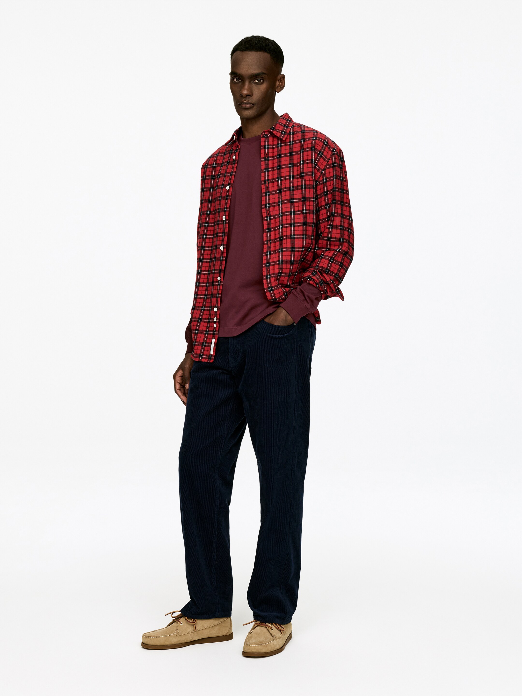 Agrandir l'image: Une chemise à carreaux rouge et noire à boutons est portée ouverte sur un t-shirt bordeaux à manches longues, les manches du t-shirt dépassant des poignets de la chemise à carreaux, tandis qu'un pantalon en velours côtelé bleu foncé présente une coupe droite, et des chaussures bateau en daim beige avec des lacets marron complètent l'ensemble. La tenue est légèrement inclinée vers la gauche, offrant une vue trois-quarts.