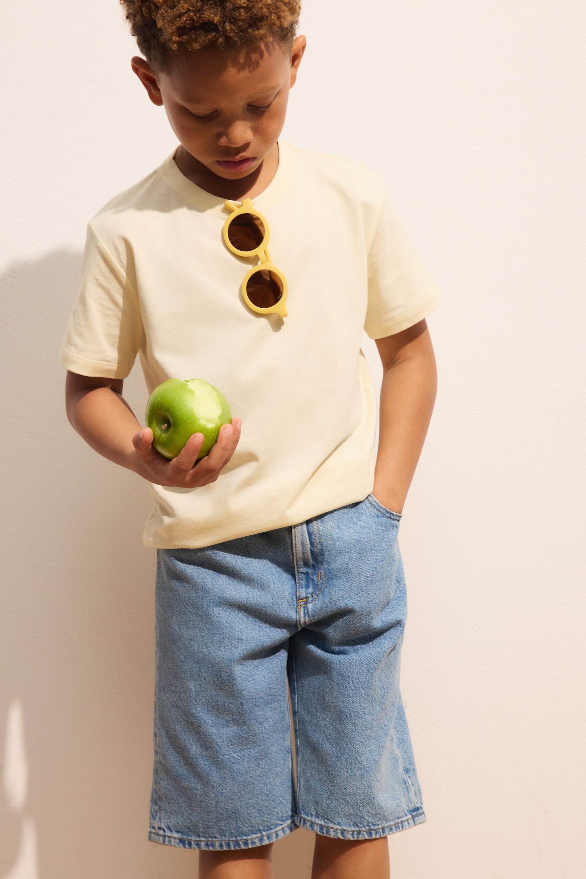 Ver imagen más grande: Se lleva puesta una camiseta de cuello redondo en color amarillo pálido, con mangas cortas y un ajuste holgado, con unas gafas de sol redondas amarillas colgando de su escote. También se llevan puestos unos pantalones cortos de mezclilla azul claro, que presentan un ajuste holgado y llegan hasta la altura de la rodilla.