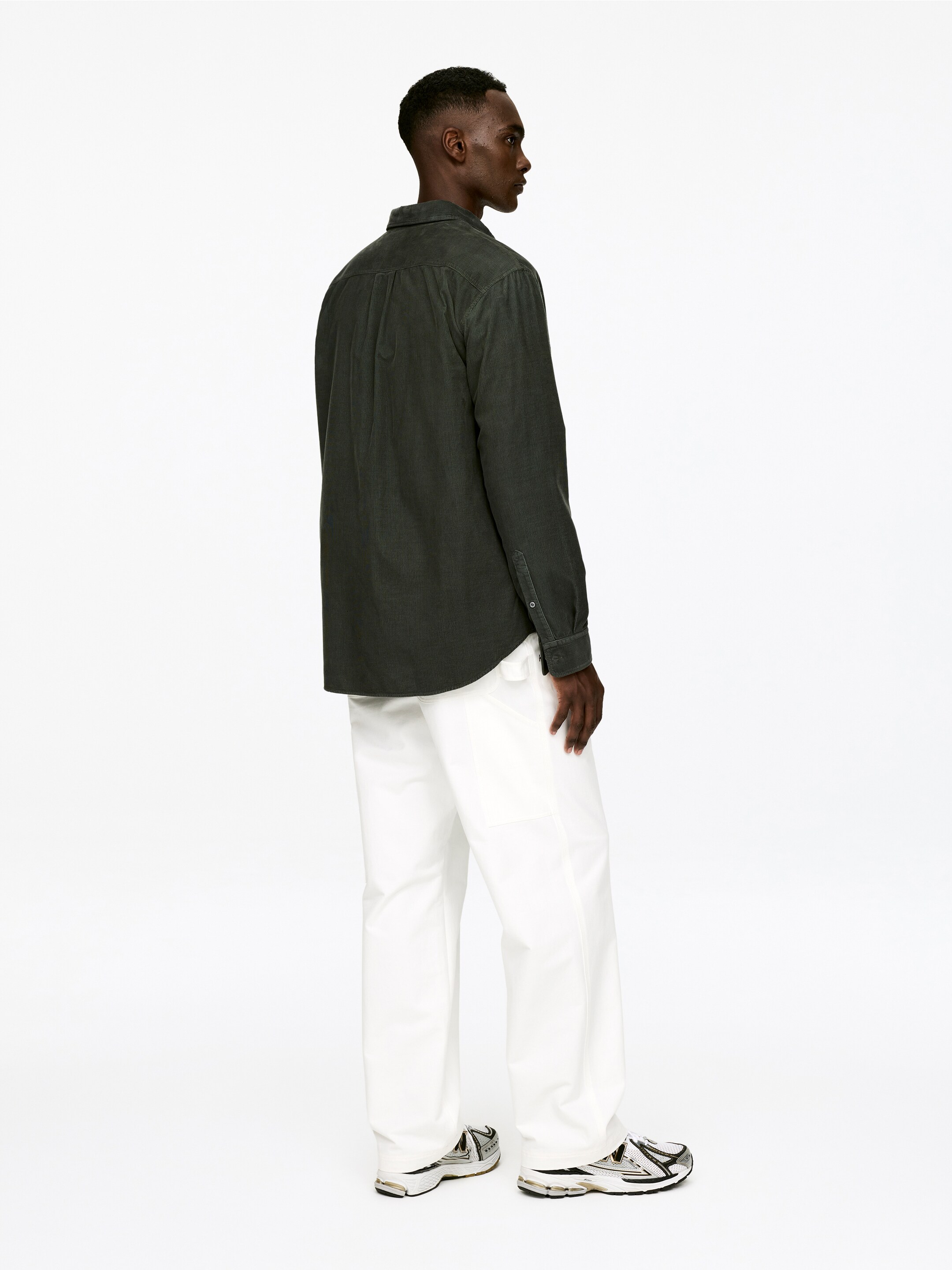 Ver imagen más grande: Una camisa de pana de canalé fino de color verde oscuro se presenta desde una vista trasera y lateral, con un ajuste holgado y puños abotonados. Actualmente se lleva desabrochada sobre unos pantalones blancos de pierna recta y holgados.