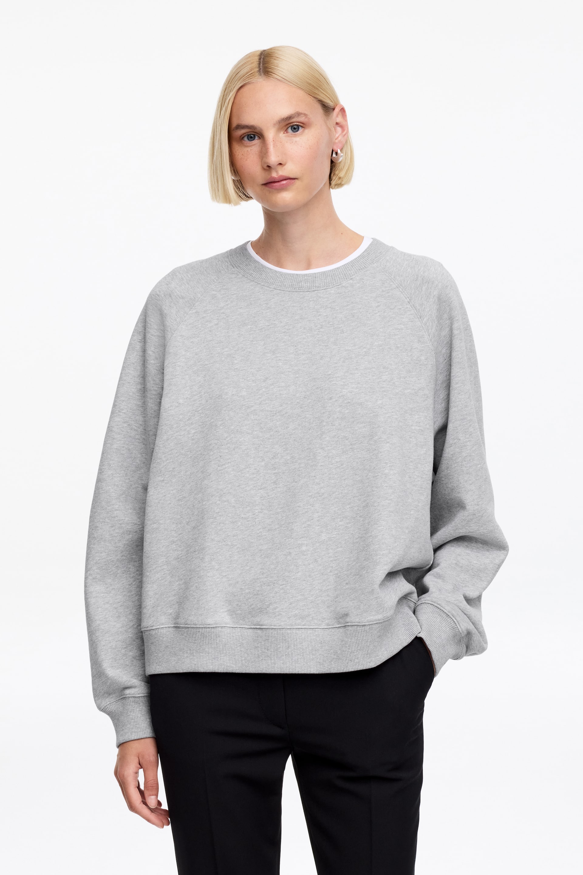Größeres Bild ansehen: Ein graues Sweatshirt wird aus einer Frontansicht gezeigt, mit Rundhalsausschnitt, Raglanärmeln, gerippten Bündchen und einem gerippten Saum. Ein weißes Rundhals-T-Shirt ist am Halsausschnitt sichtbar.