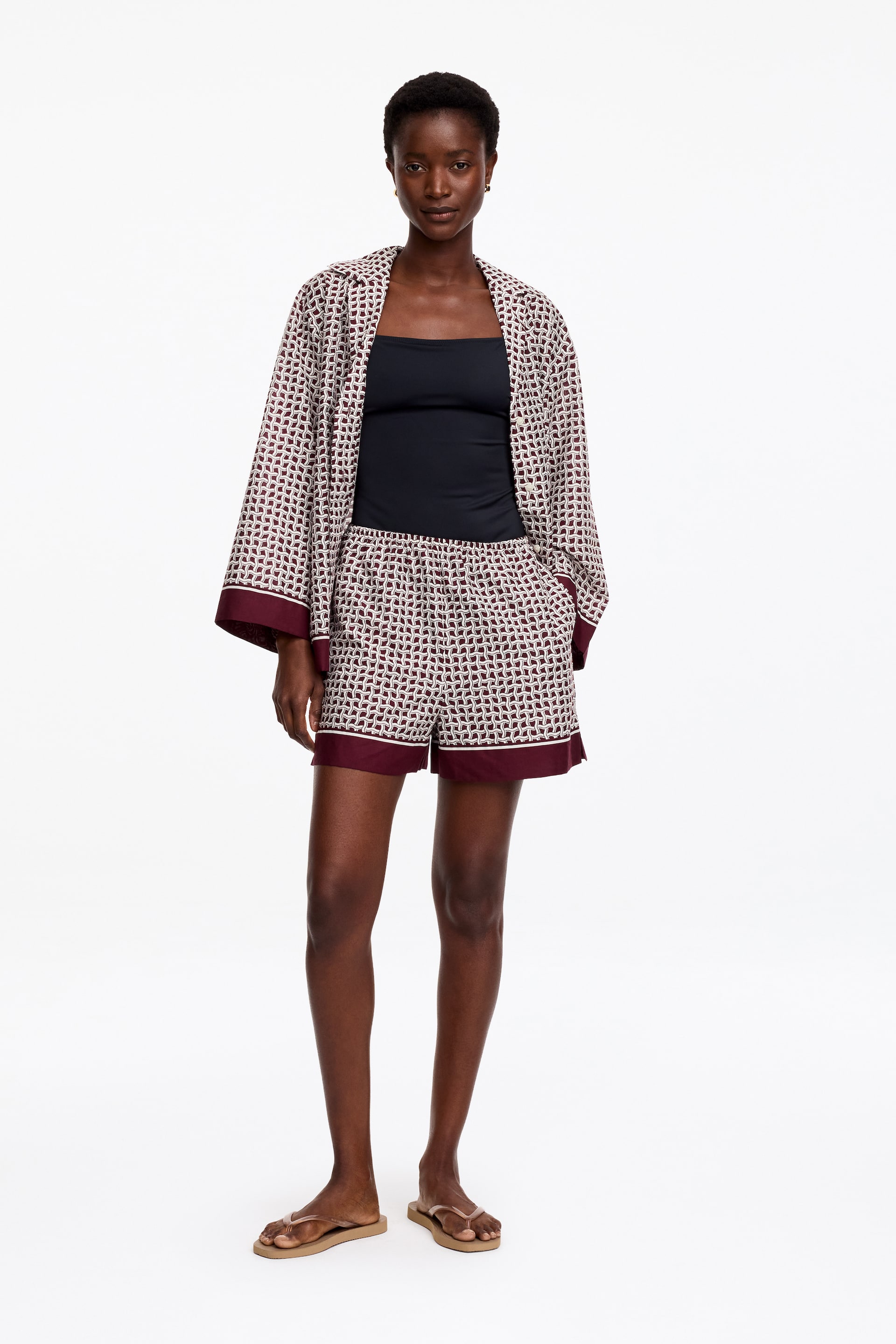 Größeres Bild anzeigen: Ein passendes Set aus einer gemusterten, offenen Jacke und hoch taillierten Shorts wird von vorne präsentiert. Die Jacke verfügt über weite, ellbogenlange Ärmel und feste kastanienbraune Manschetten, während die Shorts mit festen kastanienbraunen Säumen gestaltet sind. Beide Teile zeigen ein sich wiederholendes weißes und kastanienbraunes geometrisches Muster.