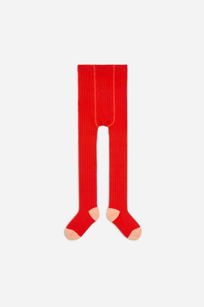 Ribbade strumpbyxor i klarrött visas framifrån, med en vertikal ribbad struktur och en elastisk midja. Modellen är detaljerad med kontrasterande häl- och tåpartier i persikofärg. - 1