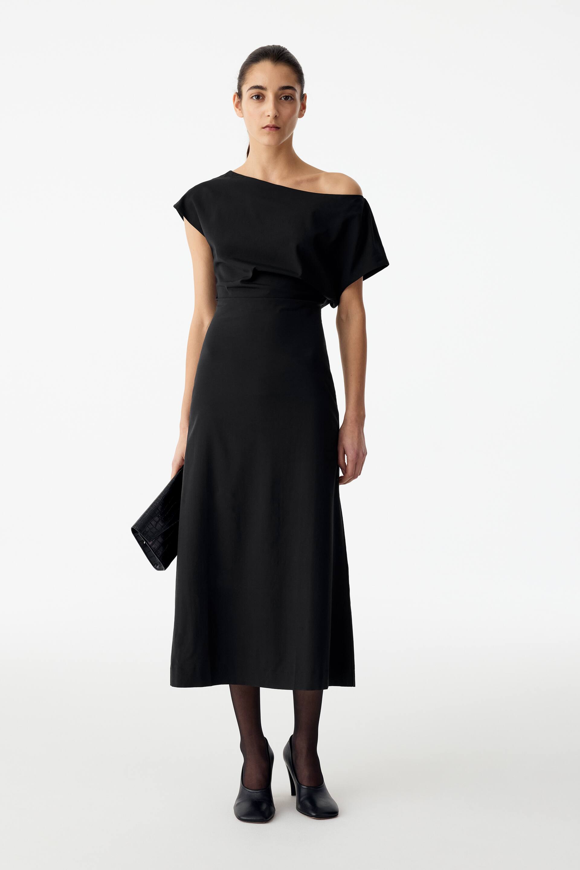 Agrandir l'image: Une robe drapée noire mi-longue, dotée d’une taille marquée et d’un décolleté asymétrique à épaules dénudées, est portée par le mannequin. La robe est stylisée avec des collants noirs et des chaussures noires à talons, tandis qu’une pochette noire est tenue à la main par le mannequin.