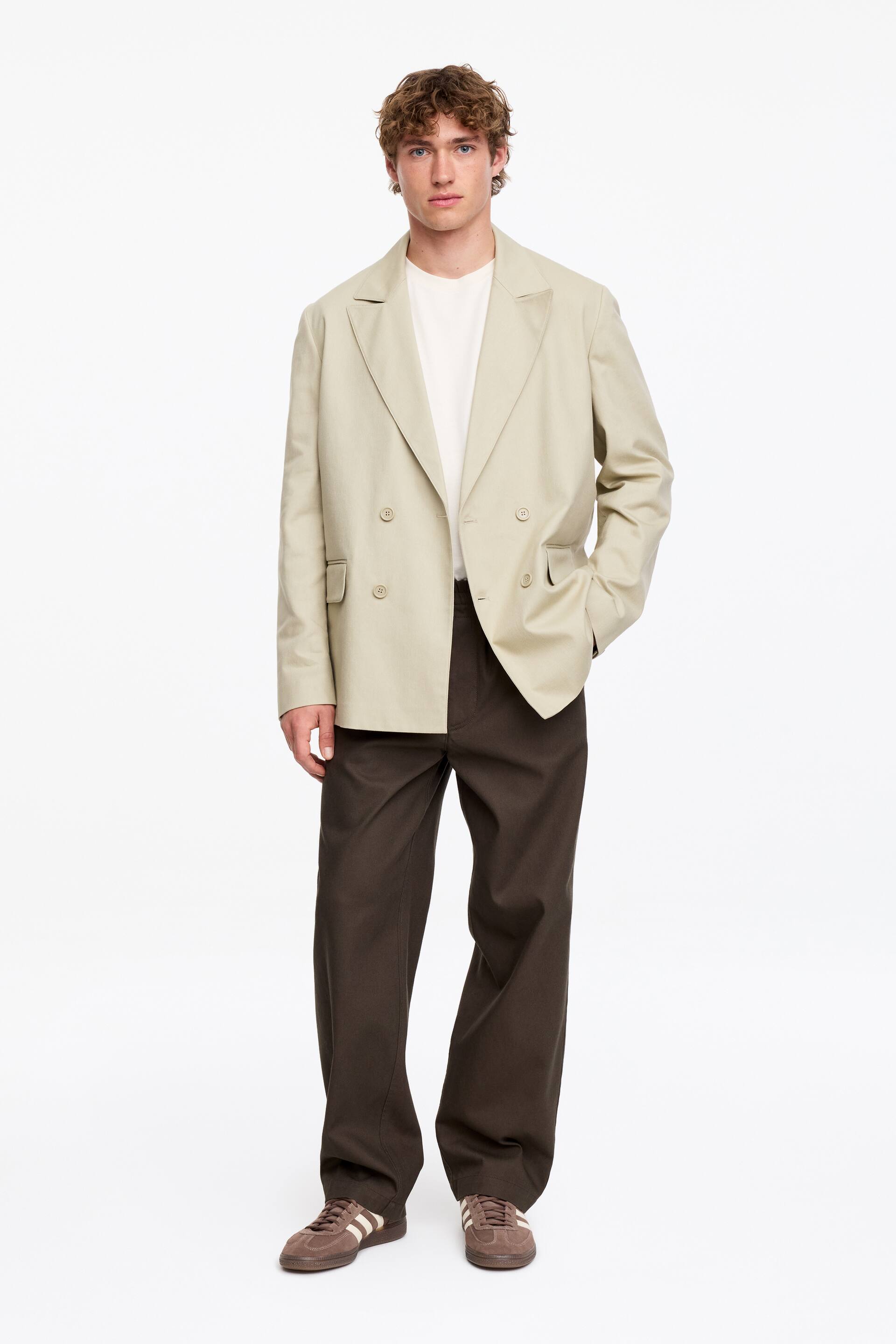 Visa större bild: En ljusbeige dubbelknäppt blazer med skårade slag och ficklock bärs öppen över en krämfärgad crew neck t-shirt. Mörkbruna byxor med vida ben kompletterar outfiten, i kombination med bruna mockasneakers med ljusa ränder och en gummisula.