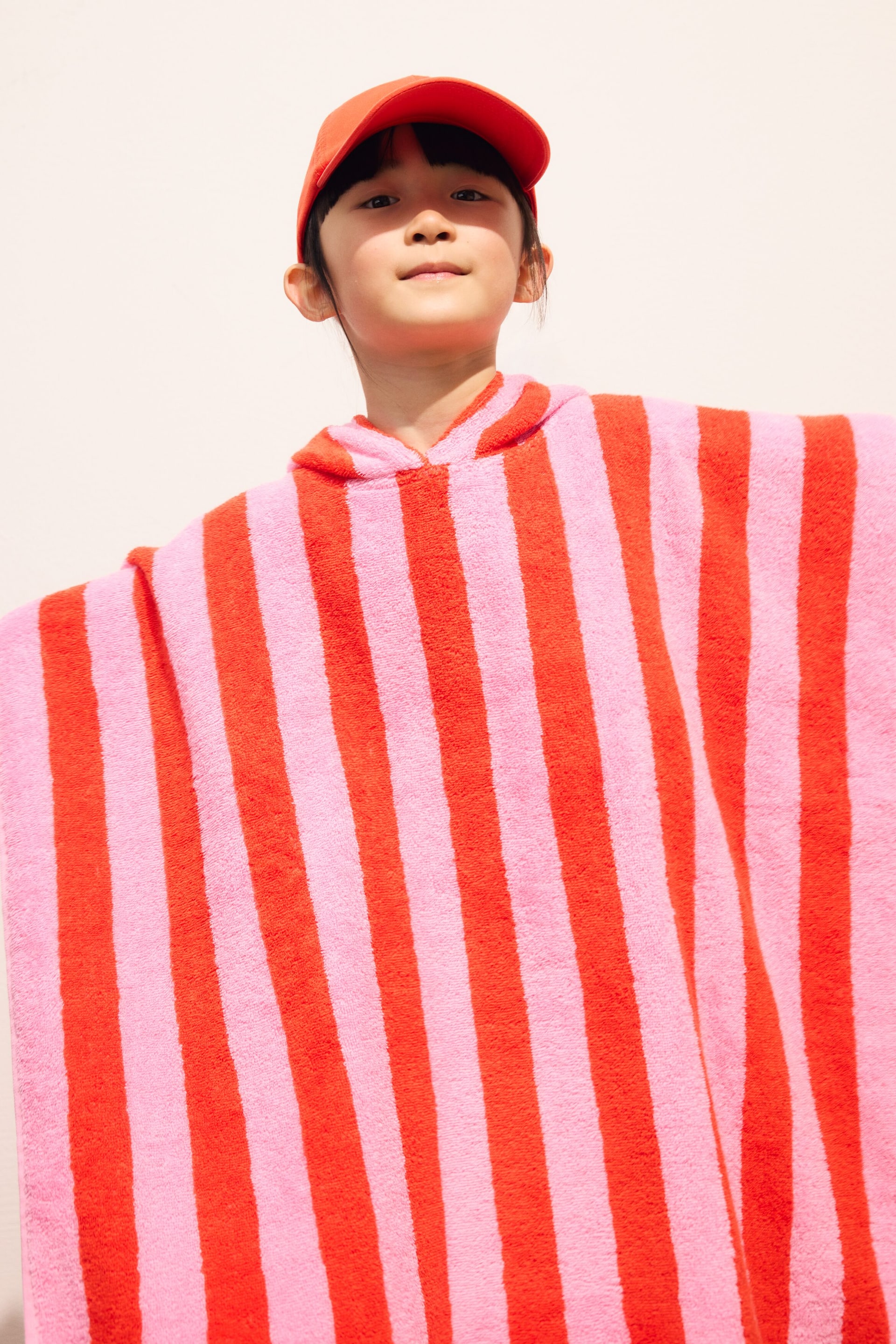 Agrandir l'image: Un poncho serviette à capuche présente de larges rayures verticales alternant entre le rose et le rouge. Il est présenté de face, avec son tissu largement déployé pour montrer sa coupe ample et sa texture en éponge.