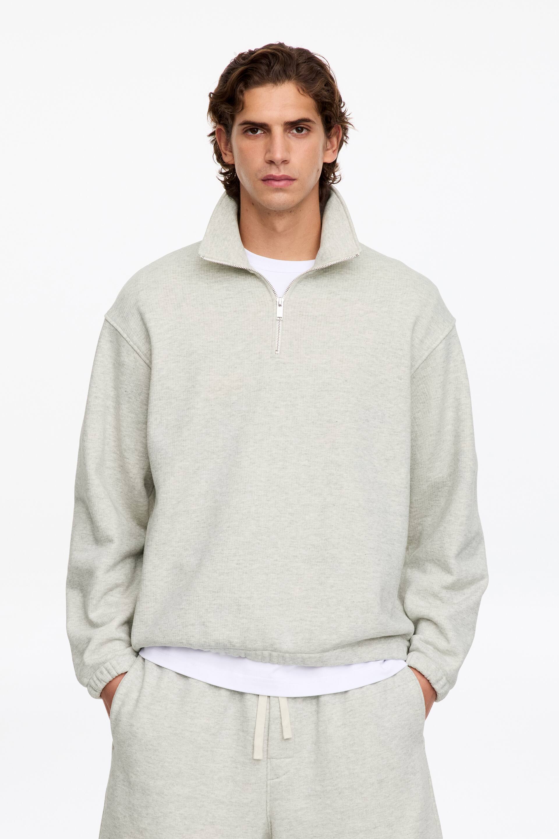 Ver imagem maior: A modelo está a usar uma sweatshirt cinzento-claro com meio fecho, gola alta, ombros descaídos e punhos e bainha elásticos. A sweatshirt está sobreposta a uma t-shirt branca e combinada com uns calções de lazer cinzento-claro a condizer.