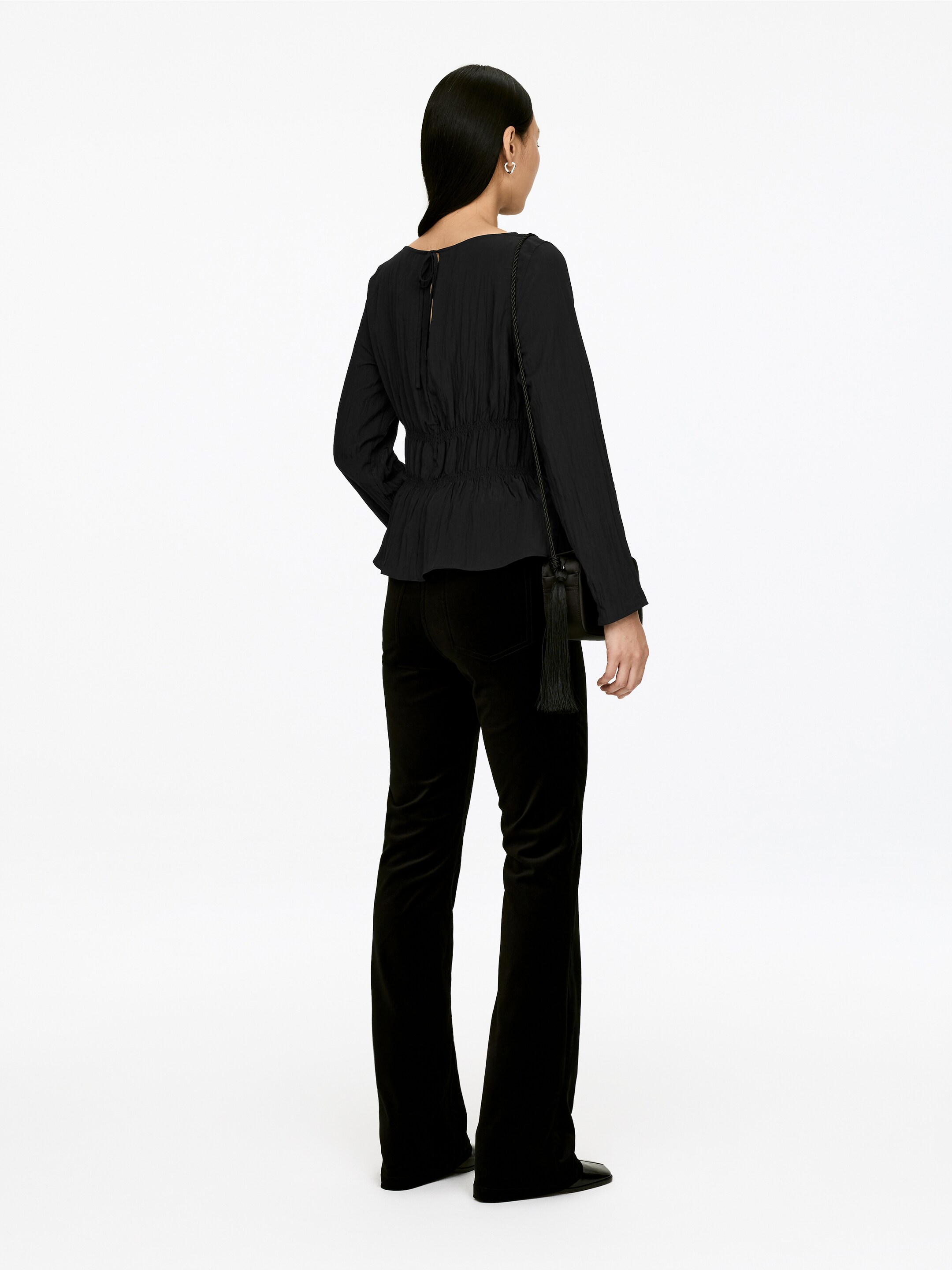 View larger image: A black crinkled long-sleeve blouse featuring a shirred peplum waist and back tie closure is being worn with black velvet flared pants, with the back of the ensemble facing the viewer.