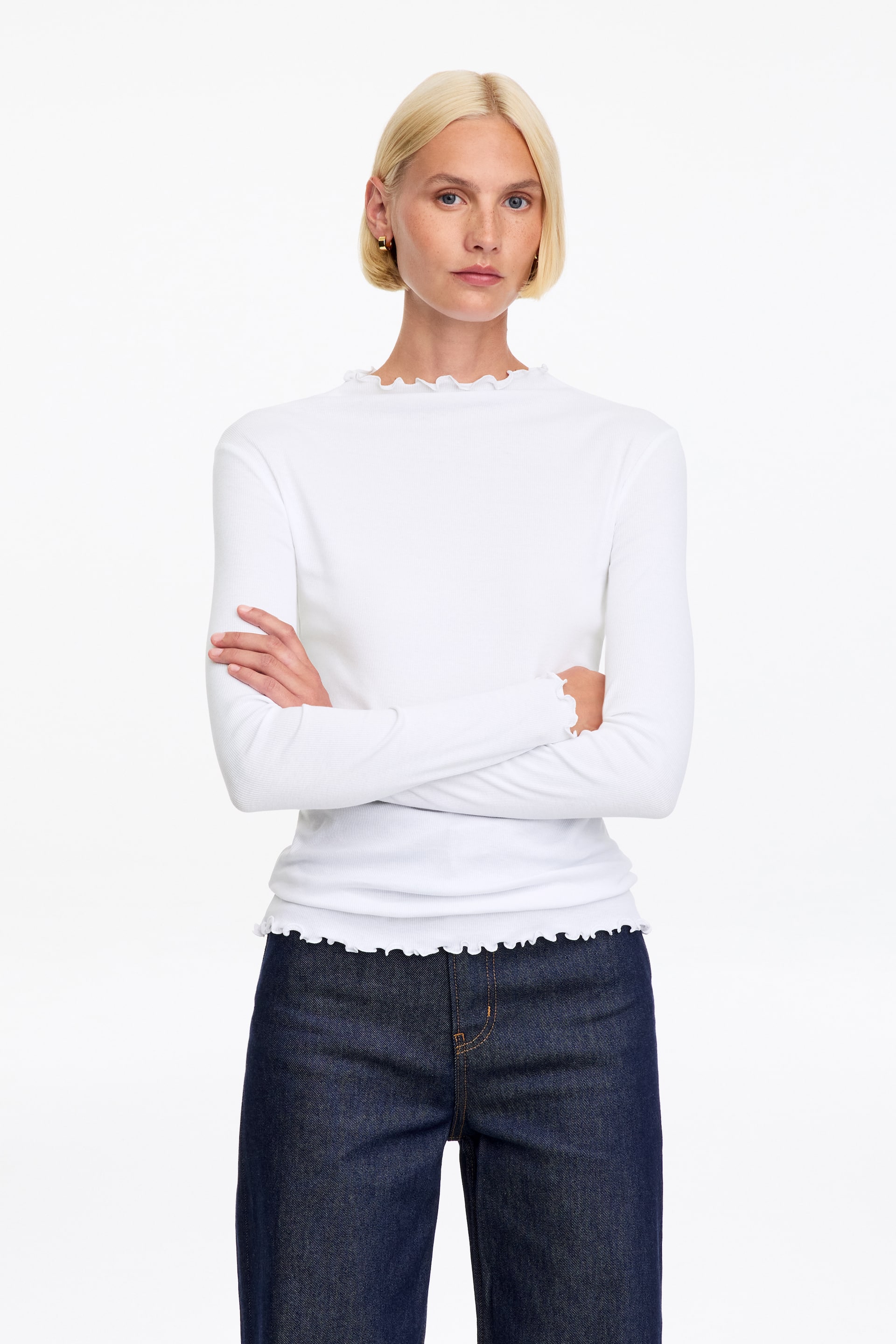 Größeres Bild anzeigen: Das Model trägt ein weißes Jersey-Top mit langen Ärmeln, hohem Ausschnitt und zarten Rüschenkanten an Kragen, Manschetten und Saum. Es steht mit verschränkten Armen und kombiniert das Top mit dunkelblauen Jeans.