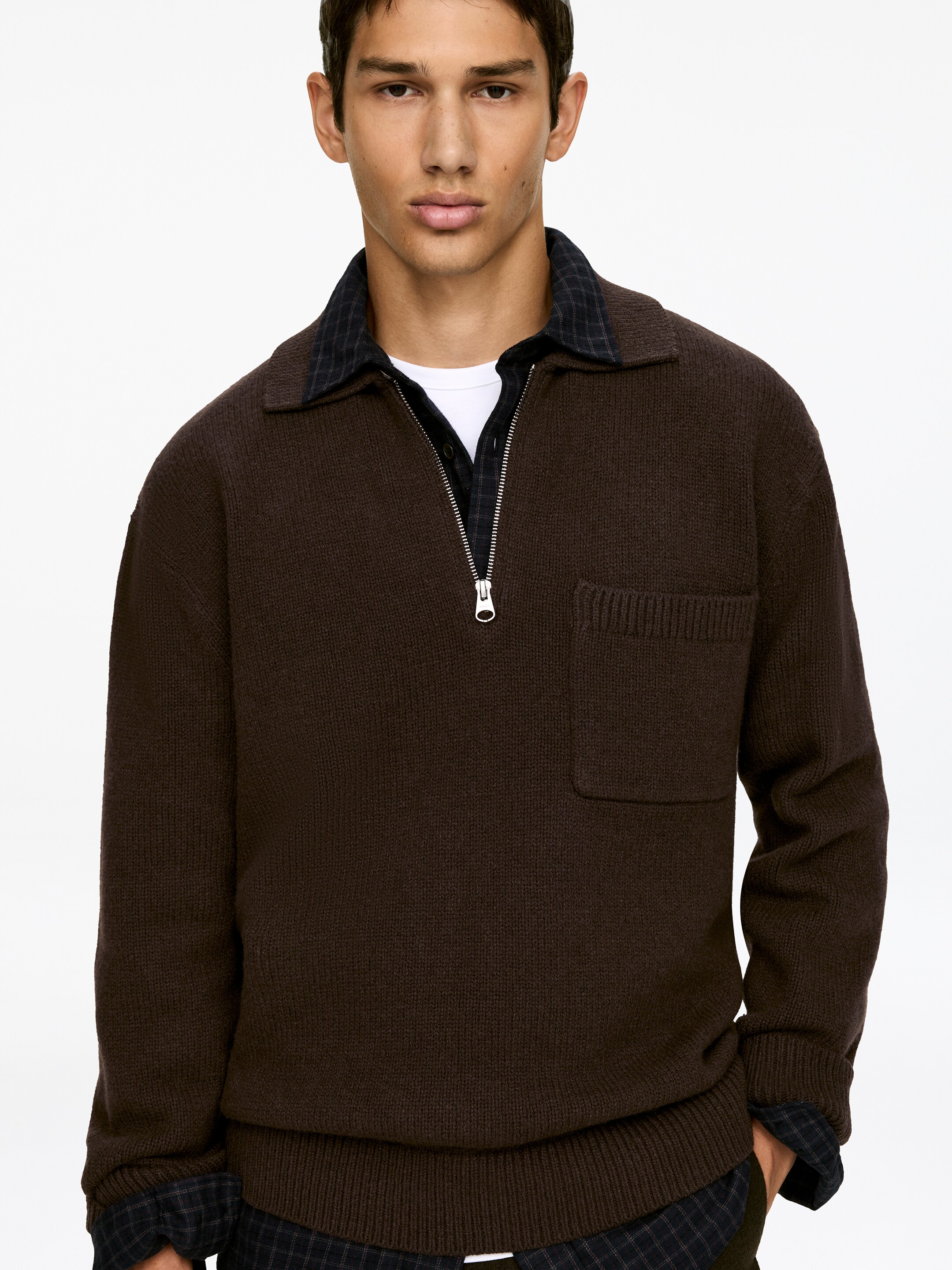 Agrandir l'image: Un pull polo tricoté marron est présenté de face, avec un col évasé, une fermeture à demi-zip avec une fermeture éclair de couleur argentée, et une poche plaquée rectangulaire sur la poitrine gauche. Un ourlet côtelé termine le bas, et une chemise à carreaux foncée est superposée en dessous, avec son col et son ourlet visibles.