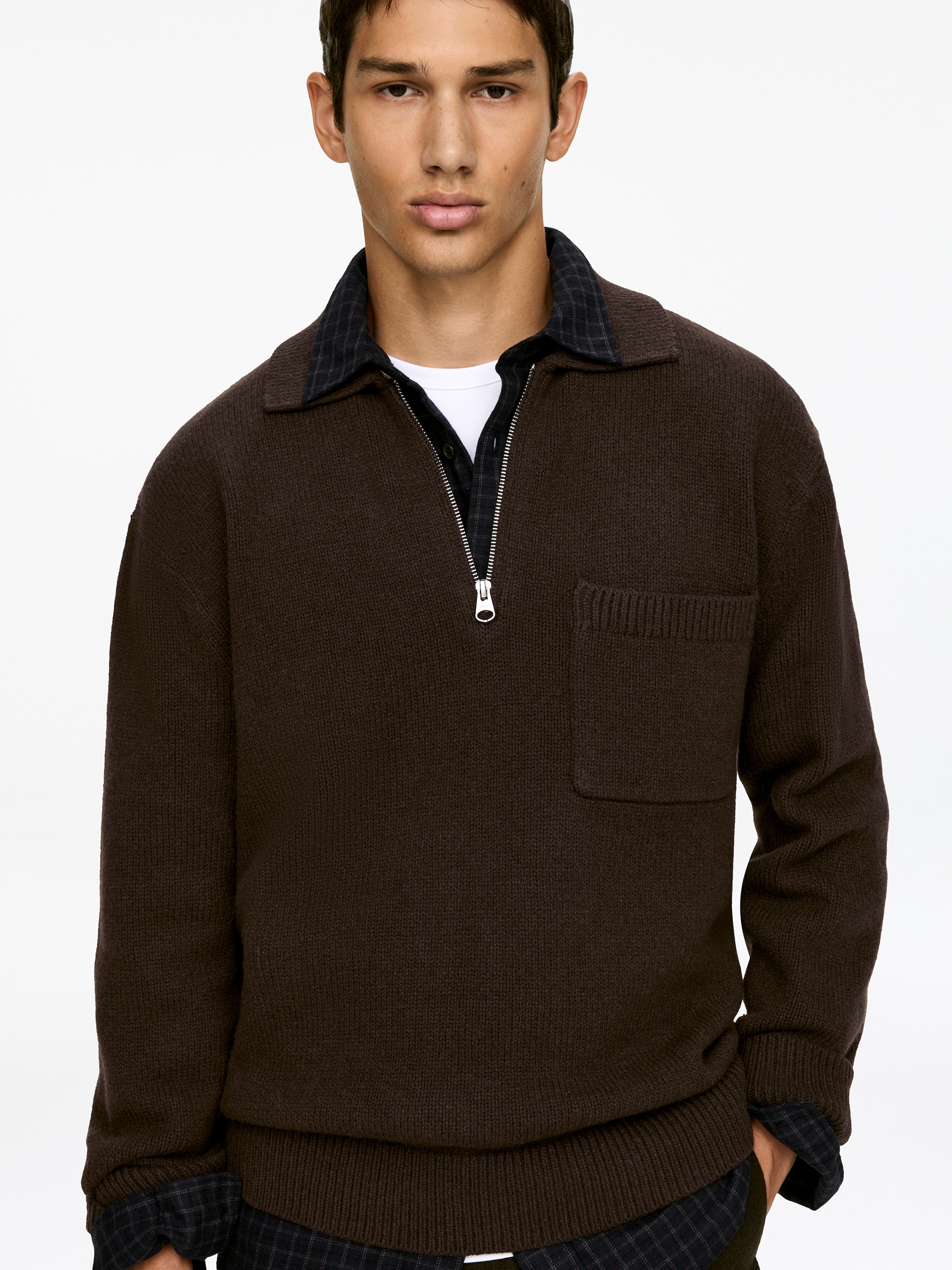 Größeres Bild anzeigen: Ein brauner Strickpolopullover wird frontal präsentiert, er verfügt über einen Kentkragen, einen Halbreißverschluss mit silberfarbenem Reißverschluss und eine rechteckige Brusttasche auf der linken Seite. Ein gerippter Saum bildet den Abschluss am unteren Ende, und ein dunkles kariertes Hemd wird darunter getragen, wobei dessen Kragen und Saum sichtbar sind.