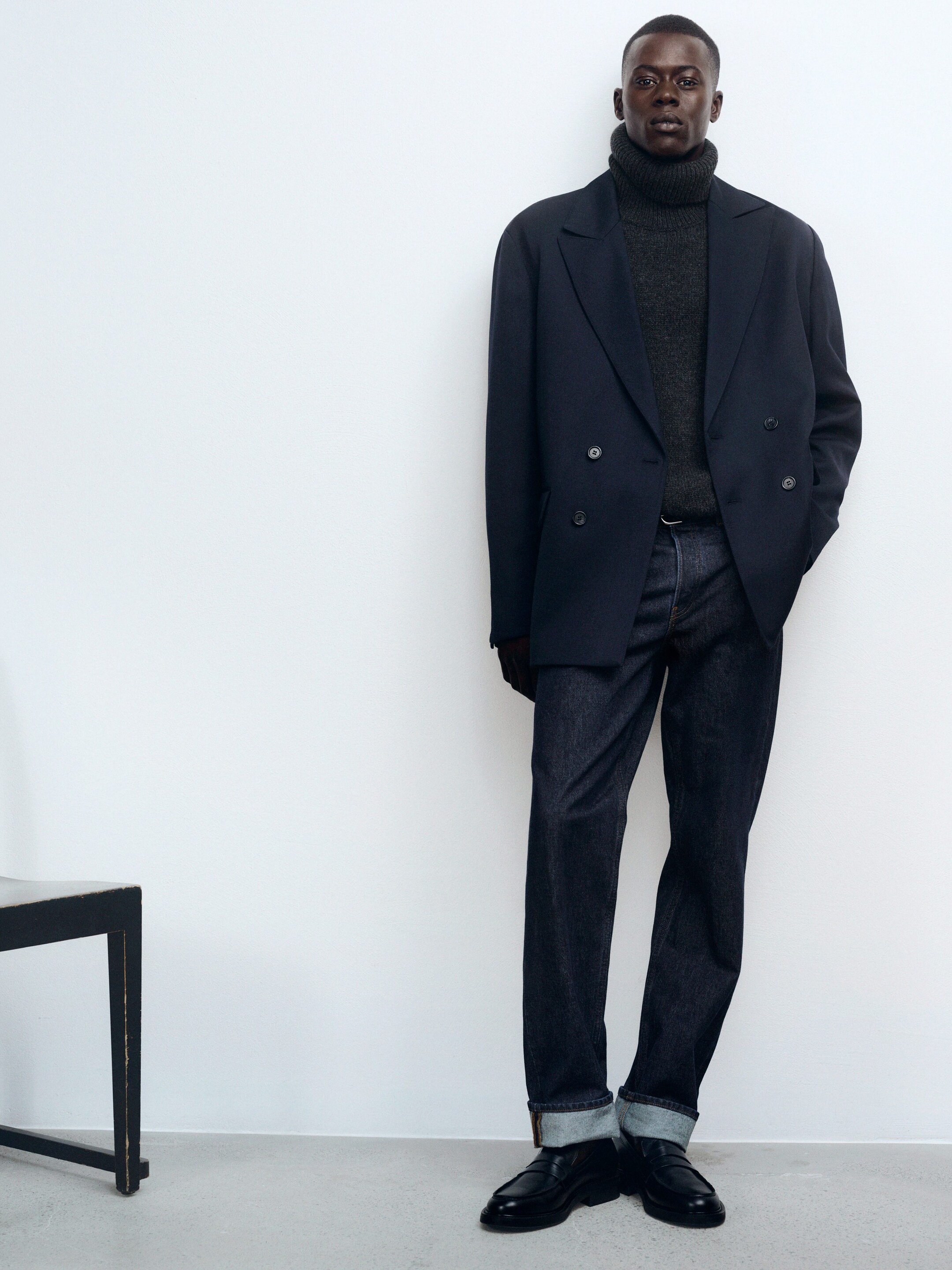 Größeres Bild anzeigen: Ein Outfit wird präsentiert, das einen marineblauen zweireihigen Blazer mit Kerbrevers und einer übergroßen, lockeren Passform zeigt, offen präsentiert und vier sichtbare Knöpfe vorne enthüllt. Ein dunkelgrauer Rollkragenpullover ist darunter geschichtet. Dunkel gewaschene, gerade geschnittene Jeans, die an den Knöcheln umgeschlagen wirken, vervollständigen den Look mit schwarzen Loafers mit klobigen Sohlen.