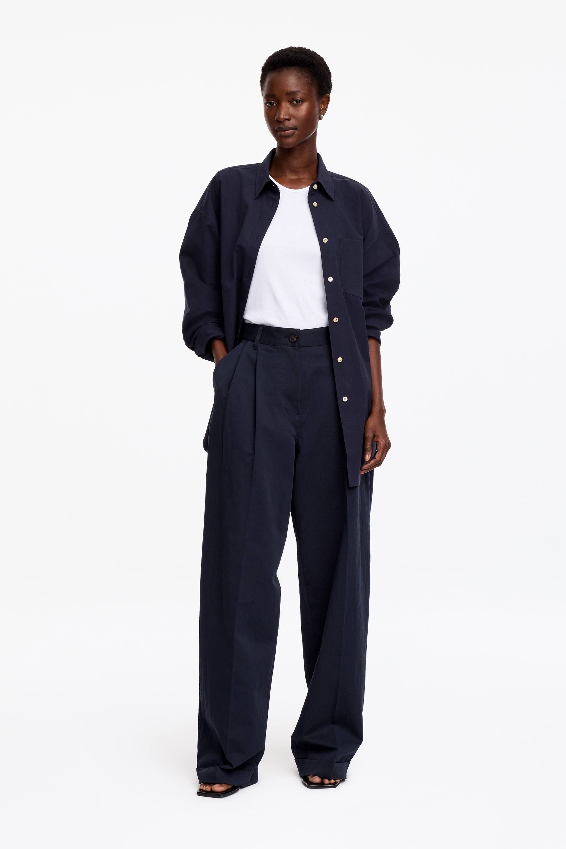 View larger image: The model is wearing navy wide-leg pleated trousers, standing with one hand in a side pocket. They are styled with an open navy shirt, a white t-shirt, and black flat sandals.