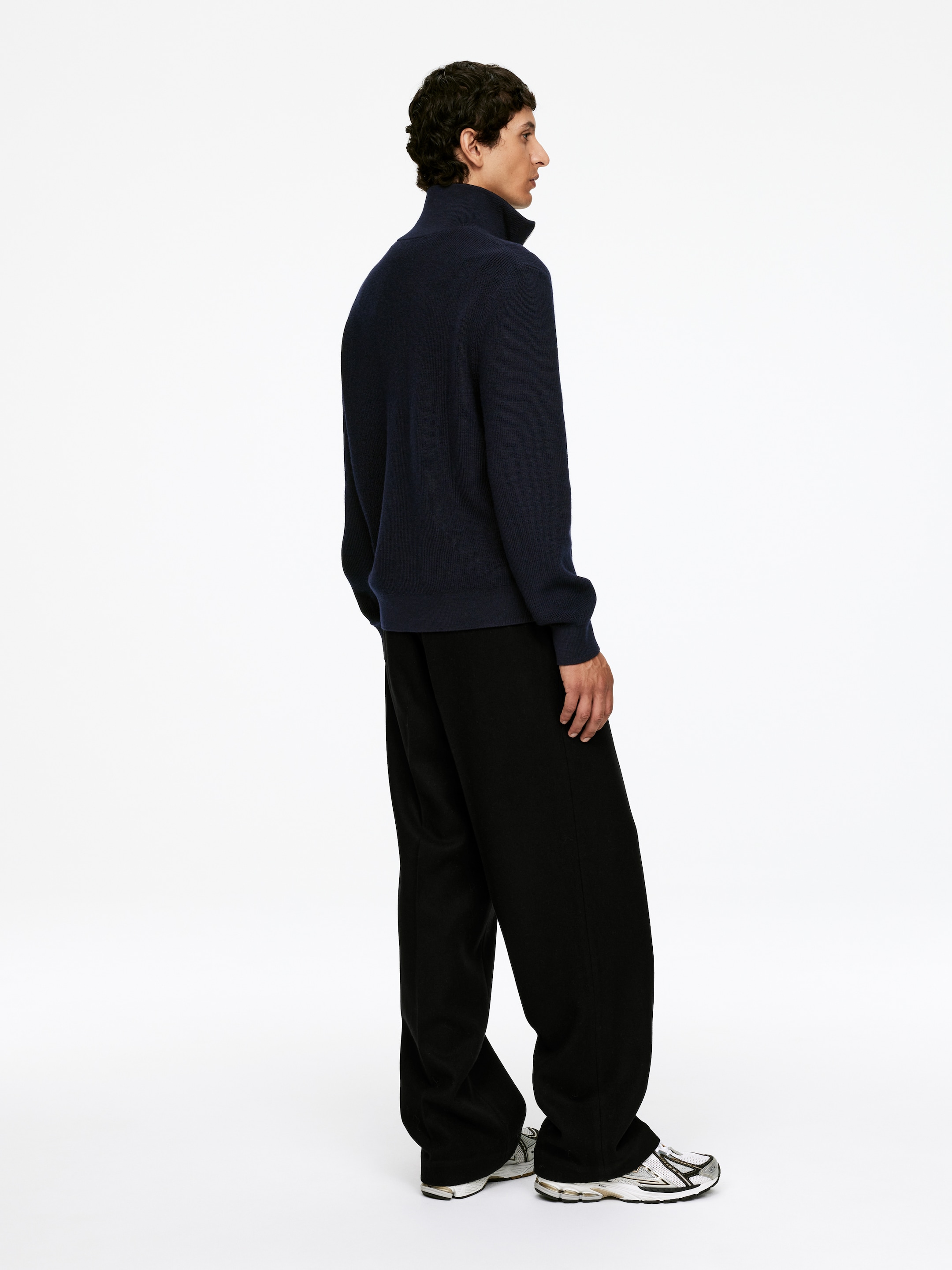 Visualizza immagine più grande: Un maglione a maglia fine blu navy viene presentato da un'angolazione posteriore-destra, caratterizzato da un colletto alto e in piedi, polsini a coste e un orlo a coste, tutti elementi che contribuiscono alla sua vestibilità rilassata. Viene abbinato a pantaloni neri a gamba larga e sneakers bianche da ginnastica.