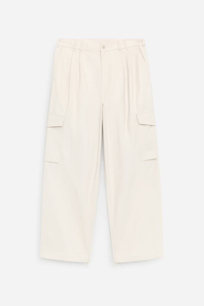 The product is a pair of cream‑coloured, loose‑fit trousers, shown from the front, featuring a button closure, deep pleats, slanted front pockets, and belt loops. Each leg features a patch pocket with a flap, and drawstrings emerge from the bottom hems. - 1