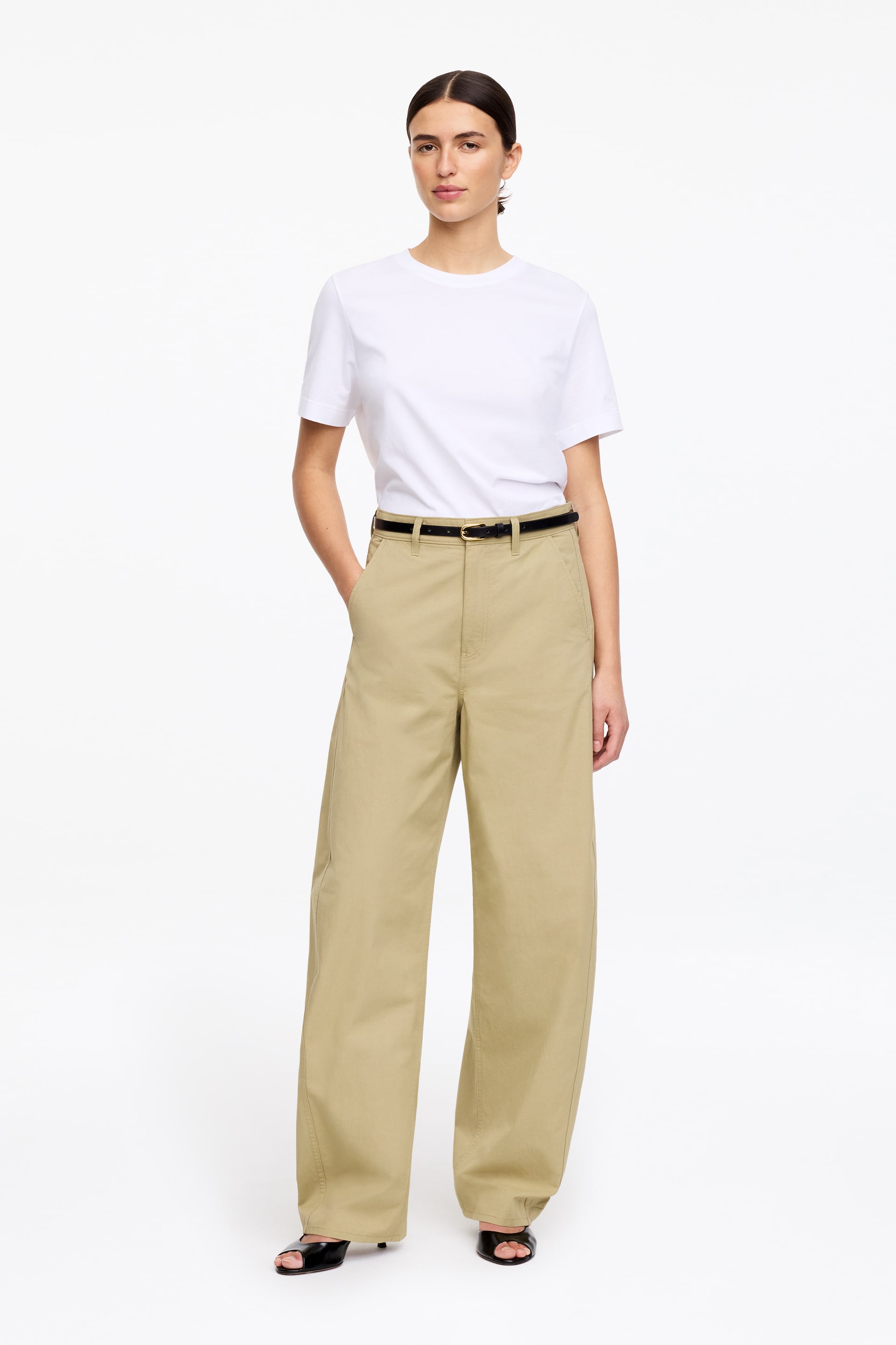 Zobraziť väčší obrázok: Biele tričko s krátkym rukávom a okrúhlym výstrihom je oblečené a zastrčené do vysokopásových, širokých khaki nohavíc, prezentované spredu, s tenkým čiernym opaskom so zlatou prackou, ktorý zvýrazňuje pás. Čierne sandále s otvorenou špičkou vykúkajú spod nohavíc.