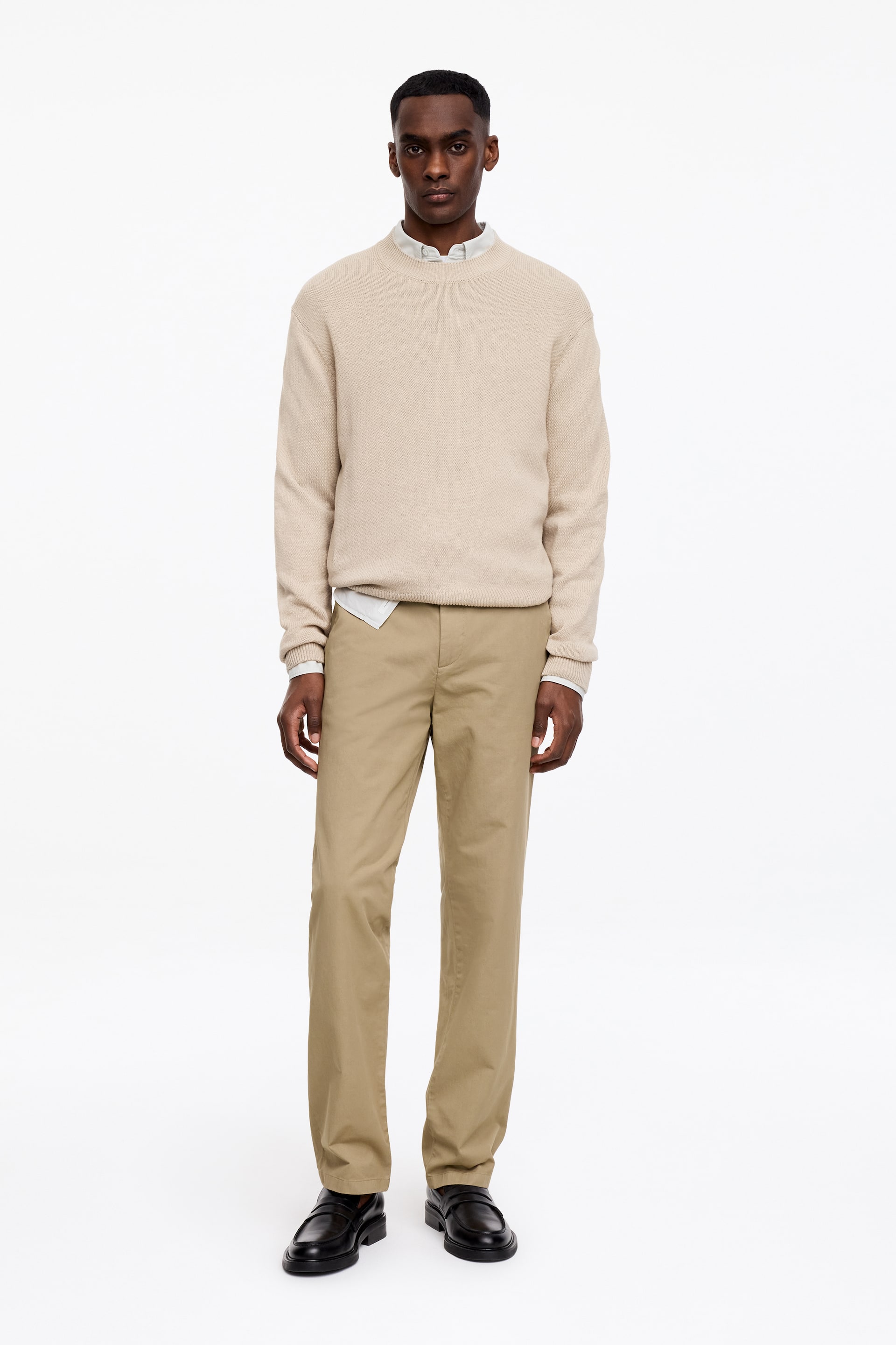 Visa större bild: En beige tröja med rund hals och texturerad stickning visas, lagrad över en ljusfärgad skjorta vars krage och nederkant är synliga. Den kombineras med khakifärgade byxor med rak passform och svarta loafers med kraftiga sulor, alla vända framåt.