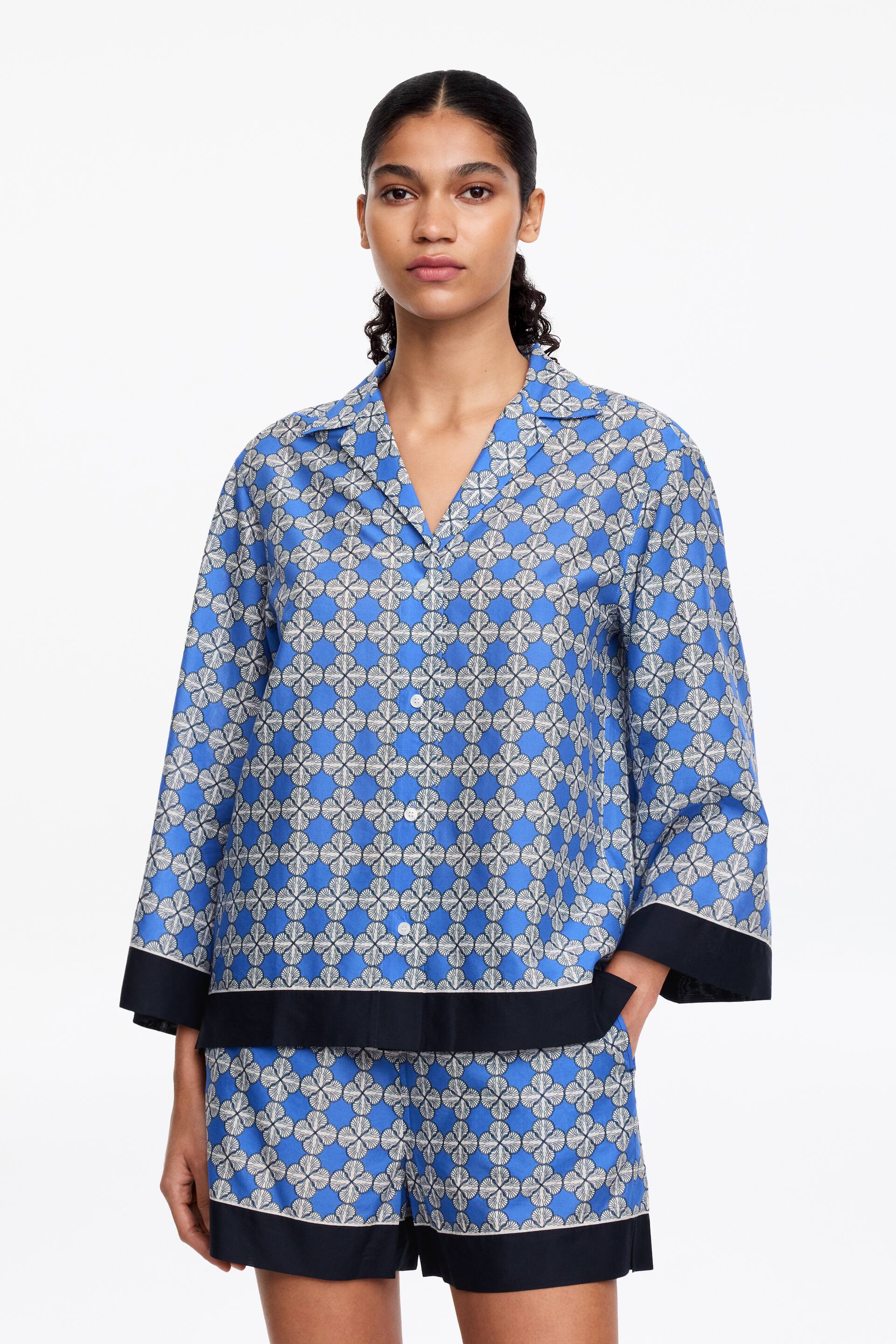 Visualizza immagine più grande: Un set pigiama blu a due pezzi presenta un motivo geometrico ripetuto in bianco e grigio. La parte superiore con colletto è progettata con un colletto a revers, uno scollo a V e maniche a 3/4, mentre sia l'orlo della parte superiore che le aperture delle gambe degli shorts sono bordati con un ampio tessuto blu navy.