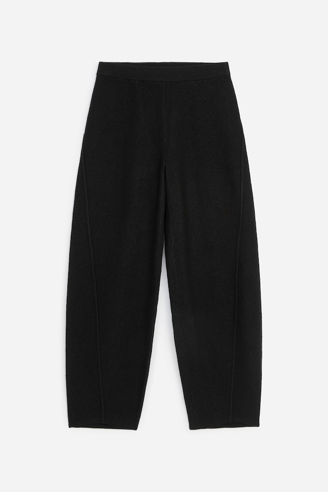 Black knit trousers are shown from the front, featuring a ribbed elasticated waistband and a relaxed, slightly tapered leg silhouette. The trousers are designed with distinct vertical seams curving along the outer sides of each leg. - 1