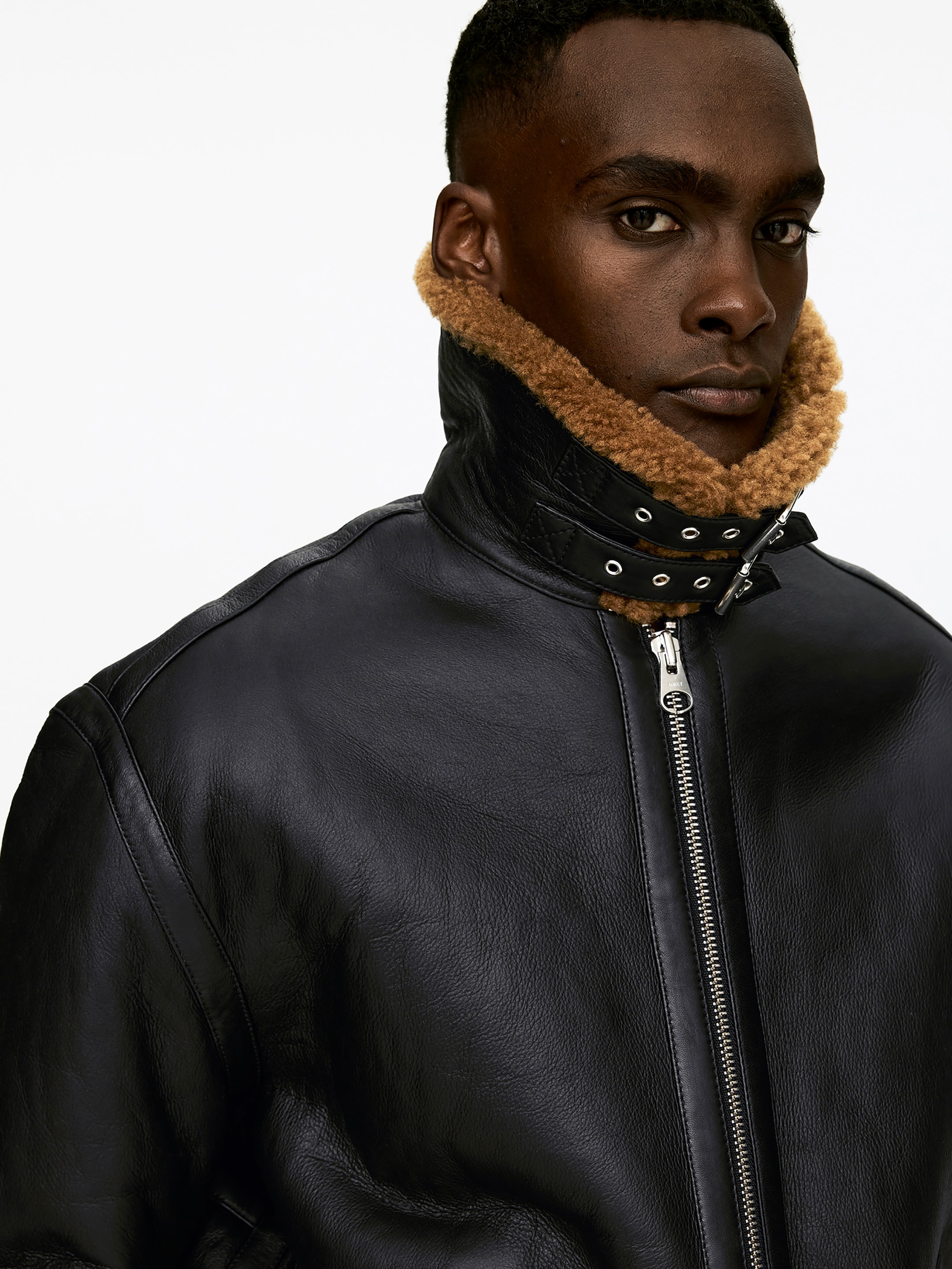 Agrandir l'image: Une veste en cuir noir présente un col montant qui est doublé de shearling marron et détaillé avec deux sangles à boucles et des œillets argentés. Une fermeture éclair de ton argenté descend au centre de la veste.