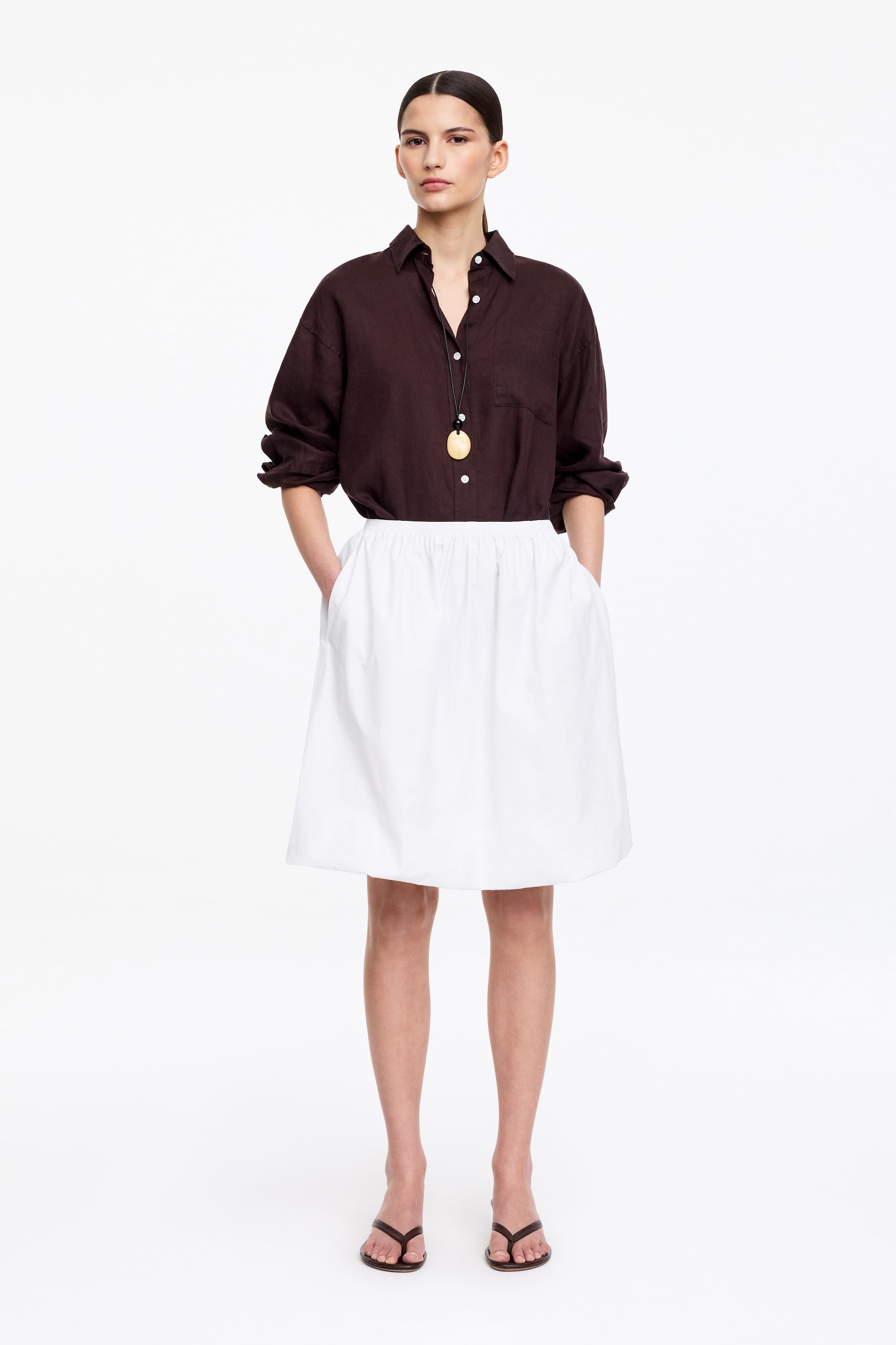 View larger image: A dark brown collared button-down shirt with white buttons and a chest pocket is being worn, its long sleeves pushed up, tucked into a white A-line skirt featuring a gathered waist and pockets. A gold-toned circular pendant on a black cord is hanging over the shirt, and dark thong sandals are being worn.
