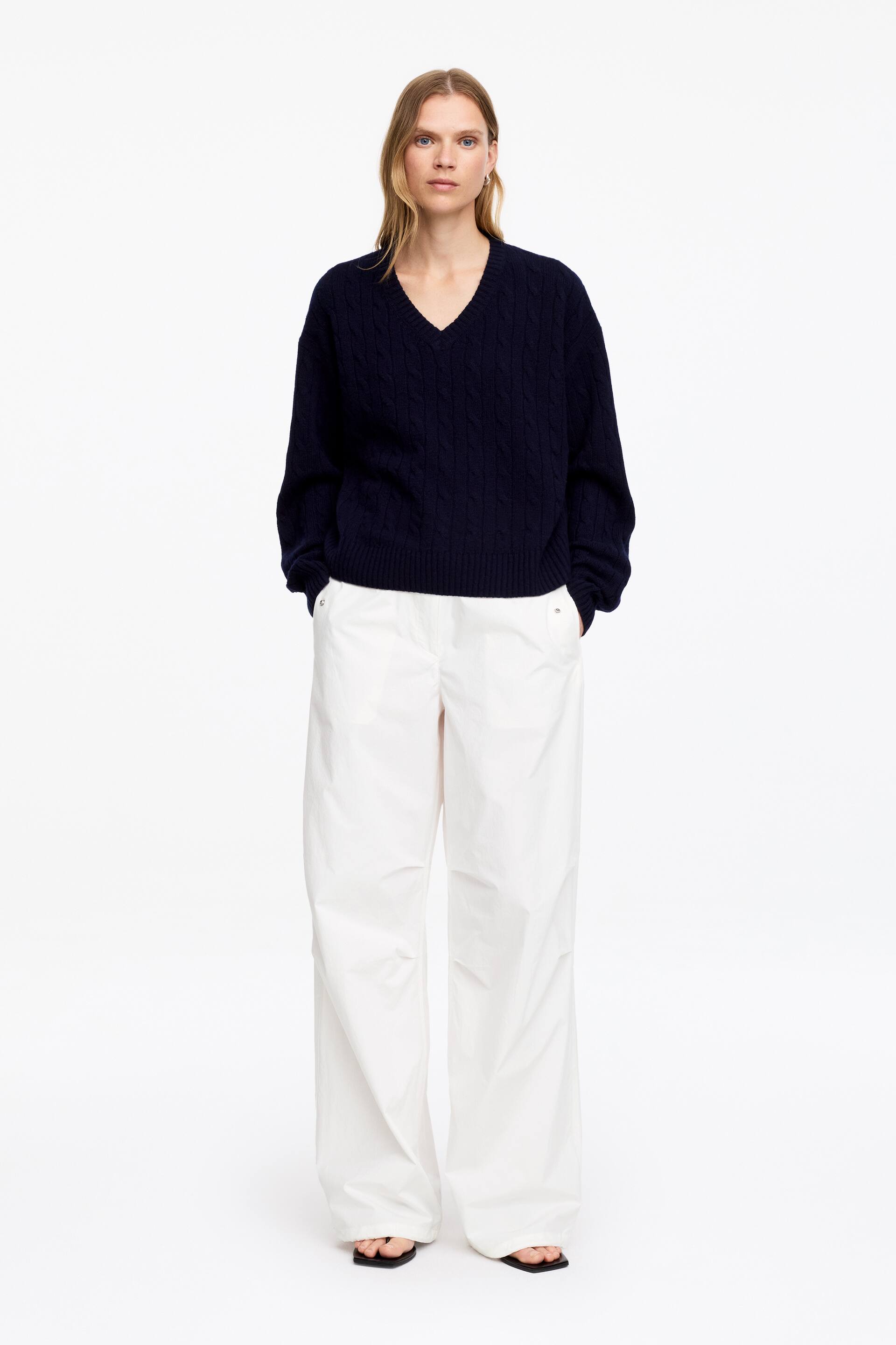 Visualizza l'immagine ingrandita: Un maglione a V in maglia intrecciata blu navy presenta una vestibilità rilassata, polsini a coste e un orlo a coste. I pantaloni bianchi a gamba larga completano il look, mostrando tasche laterali e una silhouette fluente.