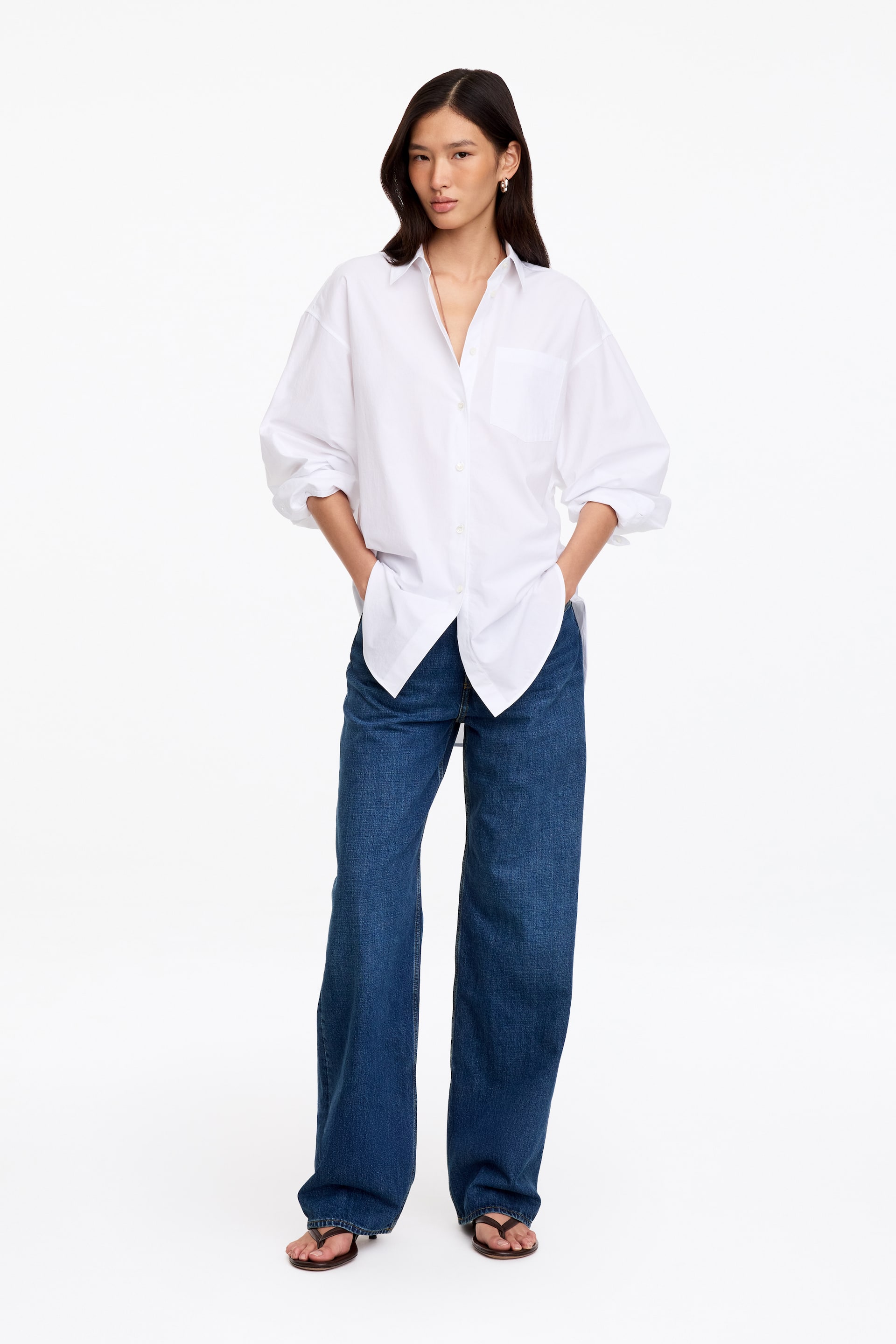 Agrandir l'image: Jean en denim bleu à jambe large et à la coupe décontractée, porté par le mannequin qui se tient debout, les mains dans les poches. Le jean est associé à une chemise blanche oversize à boutons et à des sandales tongs foncées.
