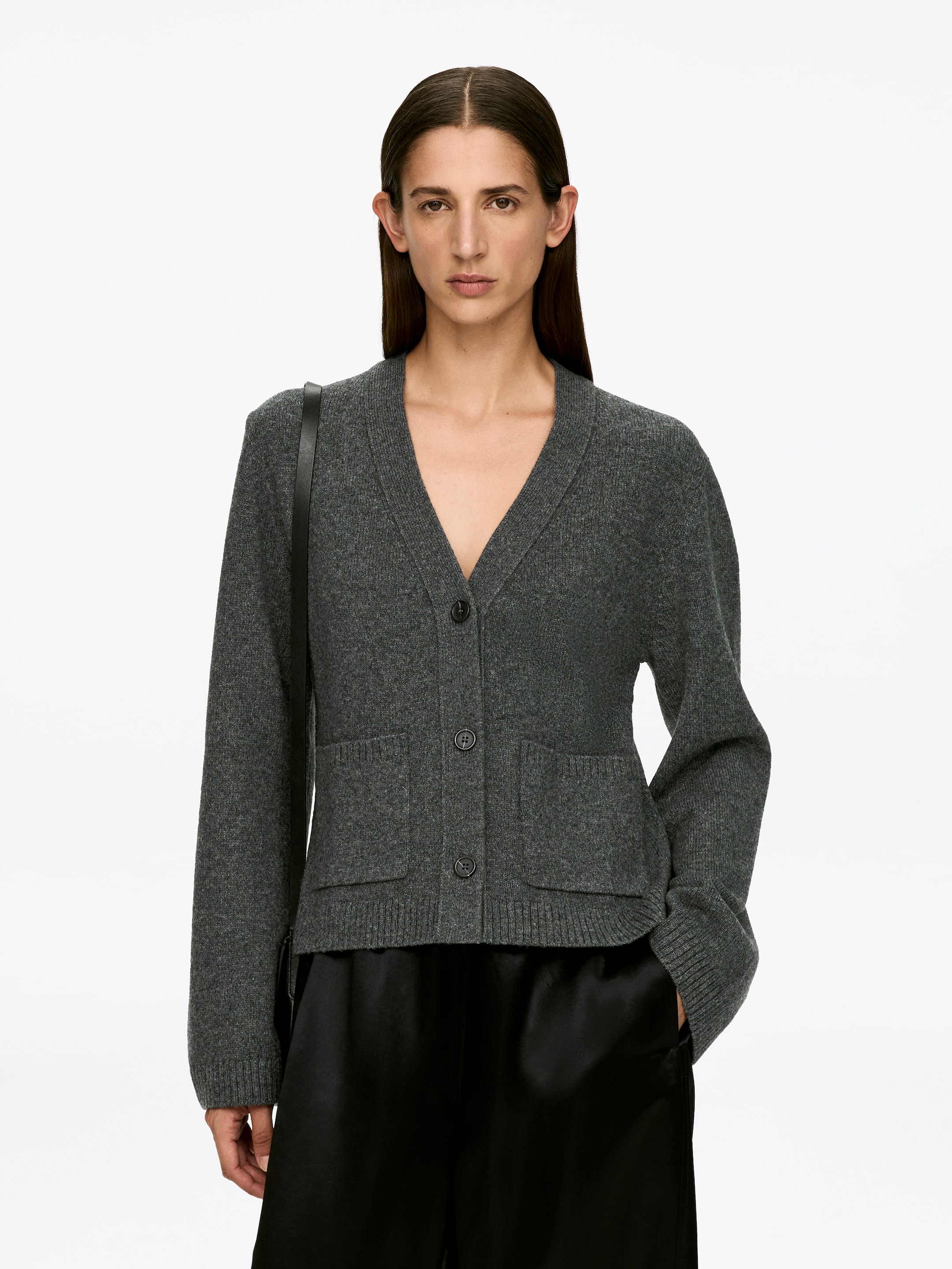 Größeres Bild ansehen: Ein dunkelgrauer Langarm-Cardigan mit V-Ausschnitt, Drei-Knopf-Verschluss und zwei aufgesetzten Taschen an der Vorderseite wird von dem Model getragen. Der Cardigan ist teilweise geknöpft, und seine schlanke Passform ist über der schwarzen Hose sichtbar.
