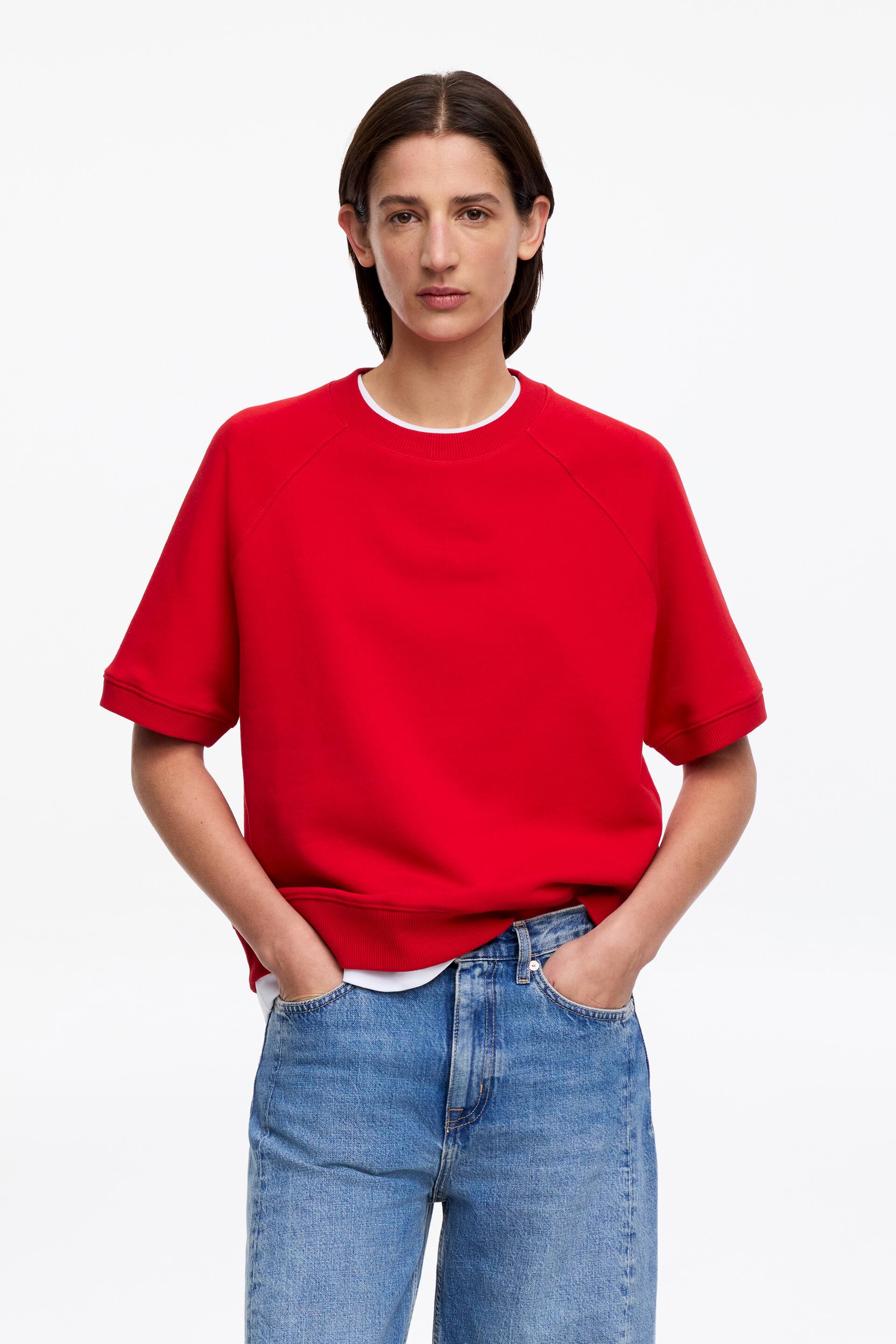 Ver imagen más grande: Una sudadera roja de manga corta presenta un corte relajado, mangas raglán, cuello redondo y dobladillo recto. Está superpuesta sobre una camiseta blanca, visible en el escote y el dobladillo.