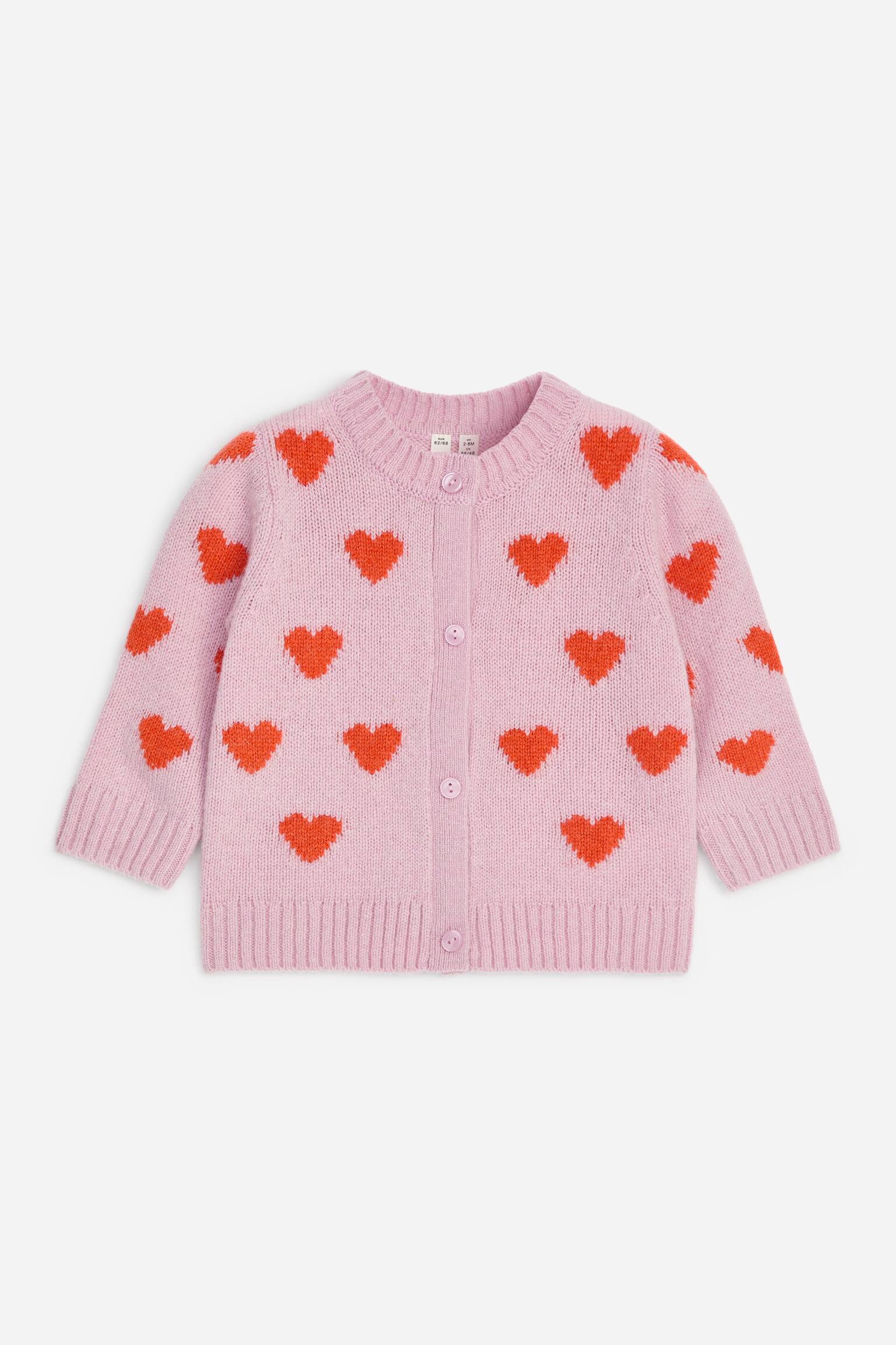 Se muestra un cÃ¡rdigan de lana rosa claro desde el frente, con un patrÃ³n integral de corazones rojos tejido en intarsia. Presenta cuello redondo, cierre frontal con botones rosas a juego y acabados acanalados en el cuello, los puÃ±os y el bajo.