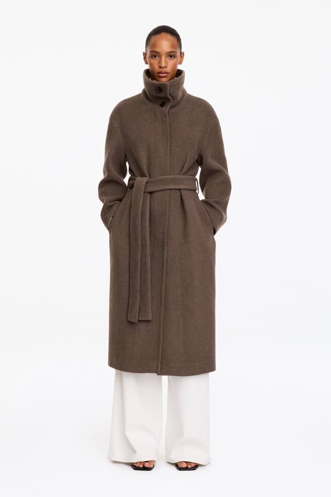 A long, brown wool-blend coat featuring a high stand collar and a matching waist-tie belt is worn by the model. The model is standing with her hands resting in the side pockets, with the coat layered over wide-leg white trousers. - 1