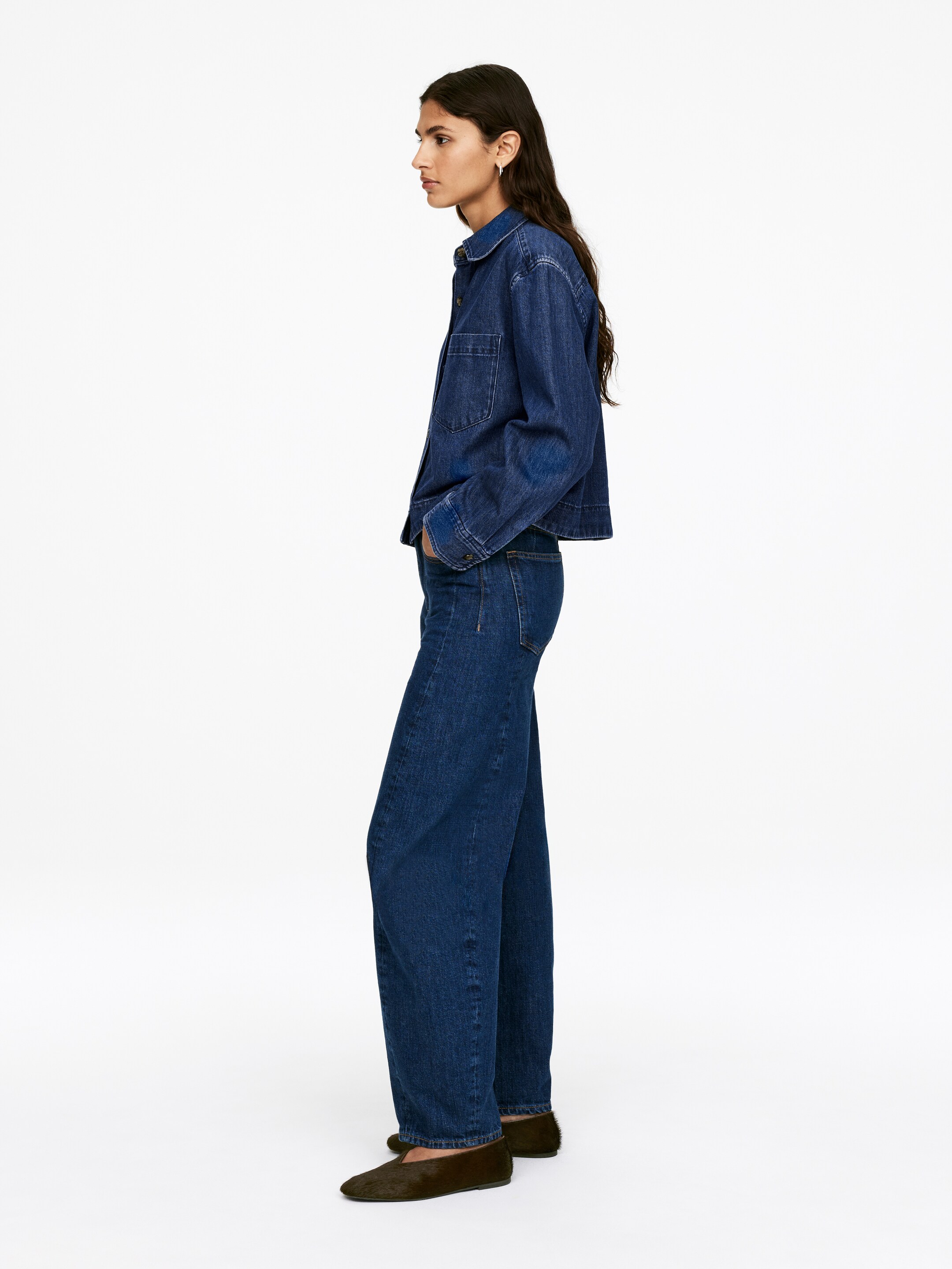 Ver imagem maior: Um conjunto de ganga de lavagem escura está a ser usado, mostrado de perfil lateral, apresentando uma camisa cropped de ajuste relaxado com colarinho pontiagudo e um bolso no peito com aba. Este top está a ser combinado com jeans de perna larga a condizer, e também estão a ser usados sapatos sem calcanhar texturizados em verde escuro.