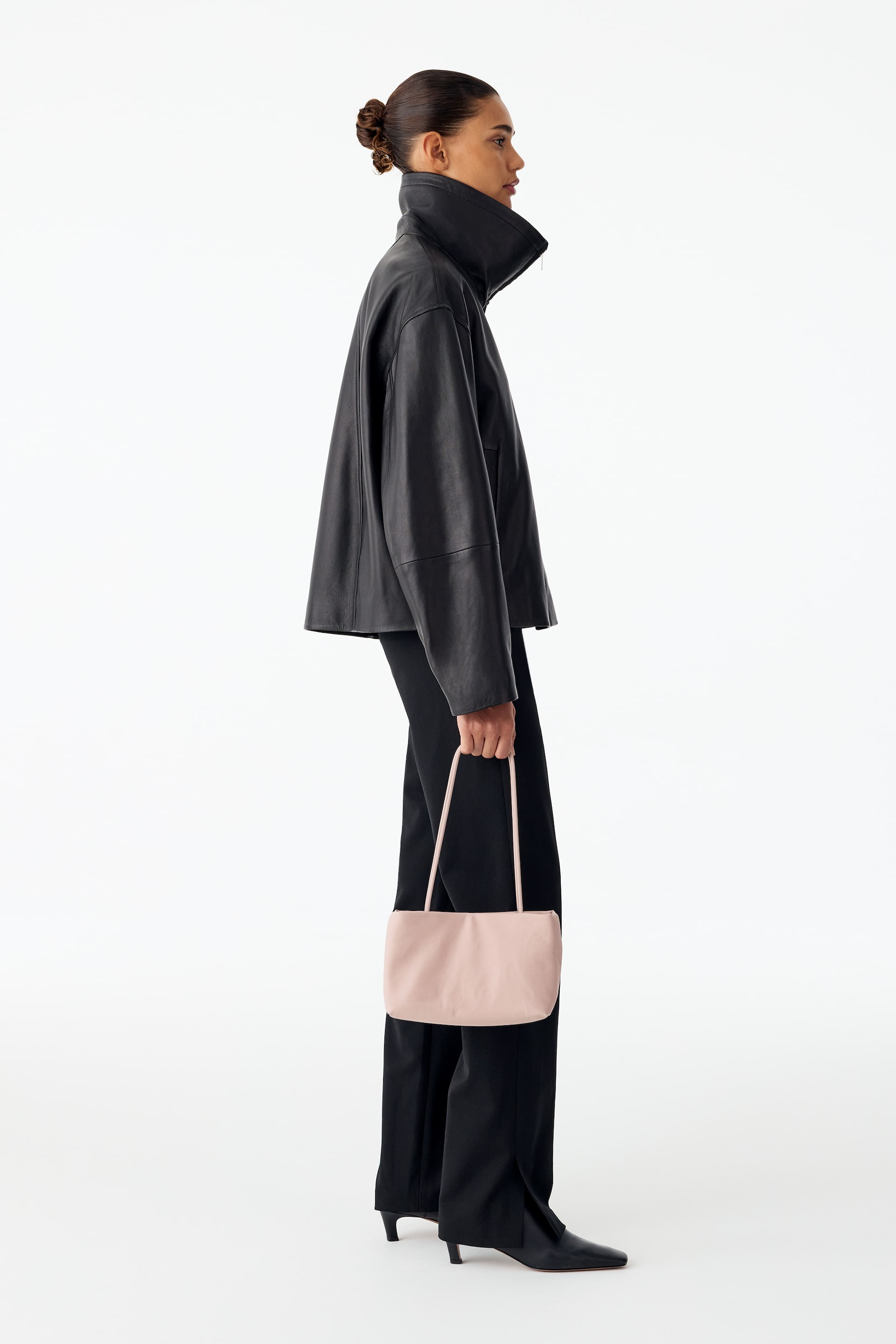 Grotere afbeelding bekijken: Een lichtroze, langwerpige boxy nylon totebag met slanke handgrepen wordt gedragen door het model, dat hem aan de hengsels vasthoudt. De tas wordt gecombineerd met een zwarte leren jas met hoge kraag en een zwarte broek.
