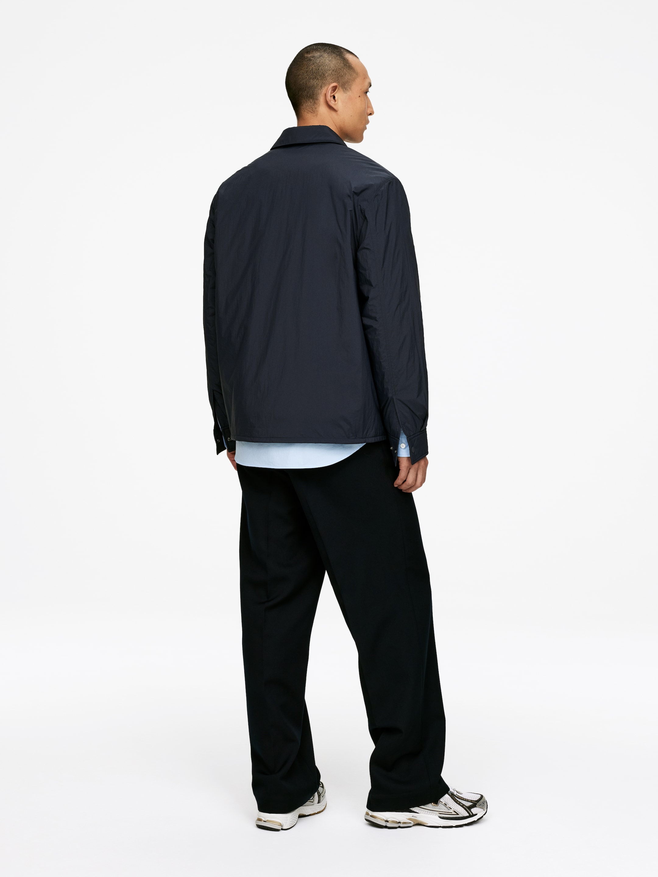 Se større billede: En mørk marineblå let overshirt, med en klassisk krave og afslappet pasform, præsenteres fra en bag-højre vinkel. Den er lagdelt over en lyseblå skjorte, synlig ved kanten og manchetterne, og er parret med mørke, brede bukser.