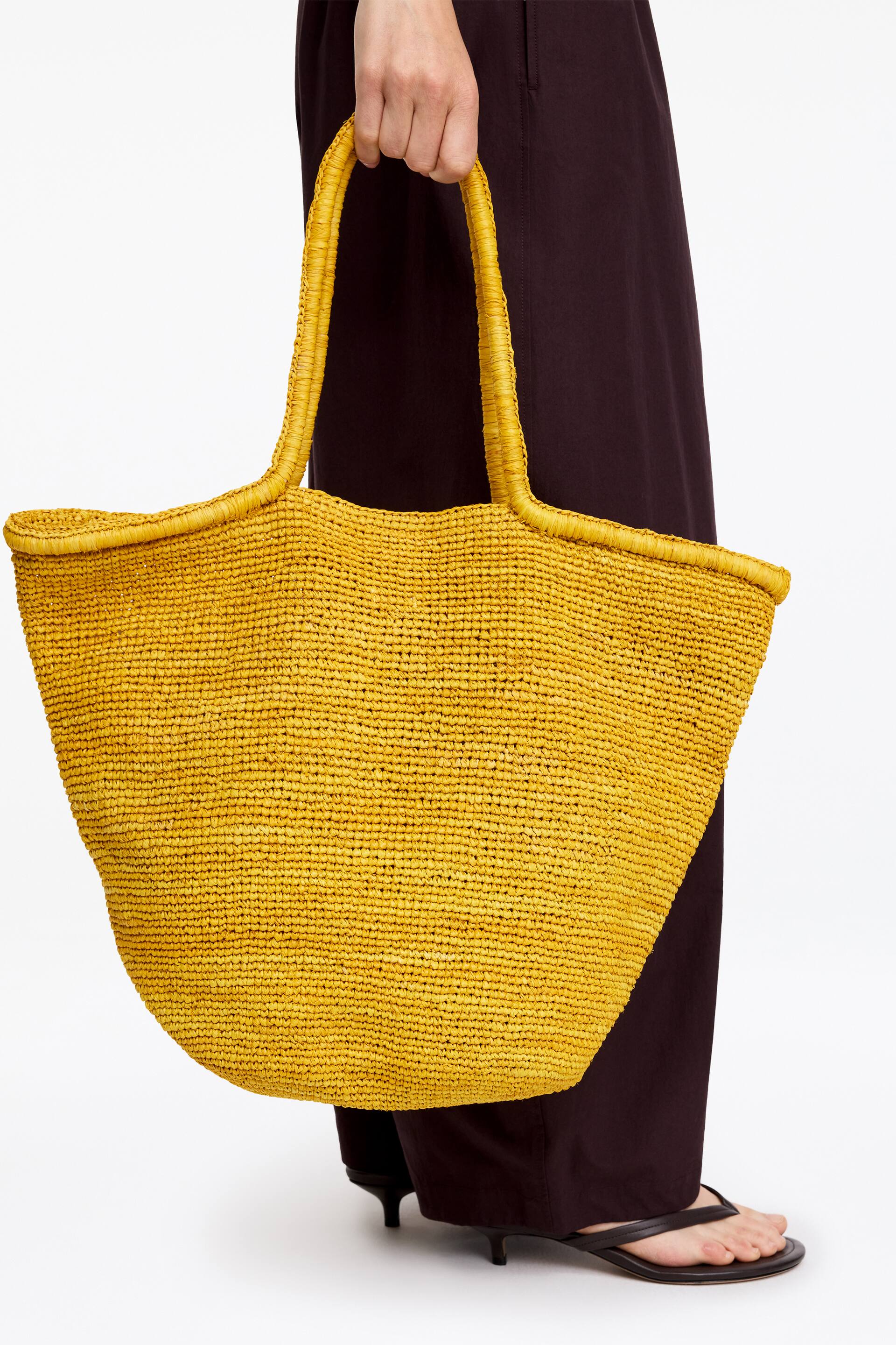Vis større bilde: En gul vevd tote-veske holdes oppreist av en hånd, som viser frem hele vesken. Den har en bred, åpen topp og to integrerte, avrundede håndtak, alt ser ut som et teksturert, raffialignende materiale.